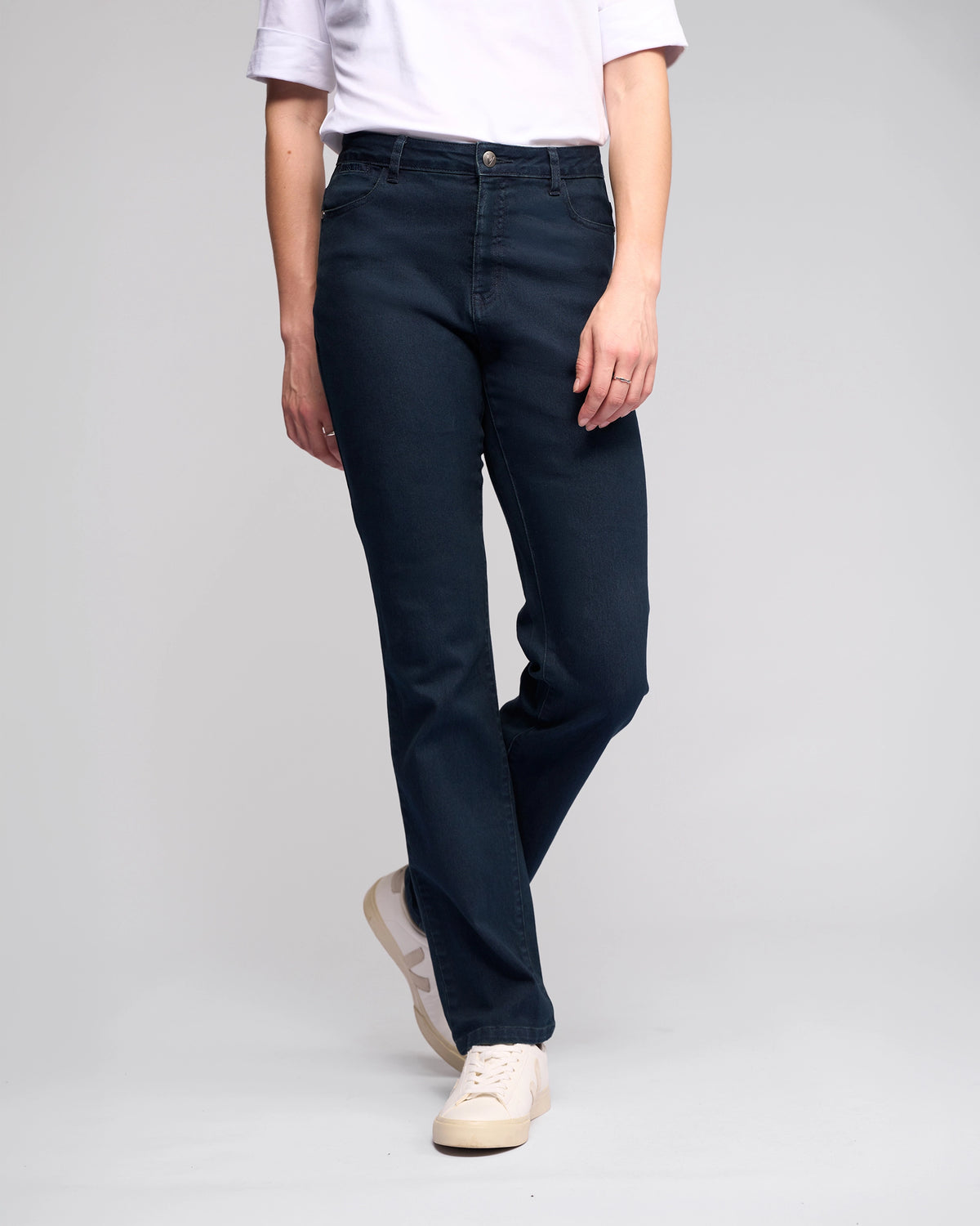 A person wears Vassalli’s Radio Jean in indigo denim, paired with a white t-shirt and white sneakers, standing against a plain light gray background. The photo captures them from shoulders to feet, arms relaxed by their sides.