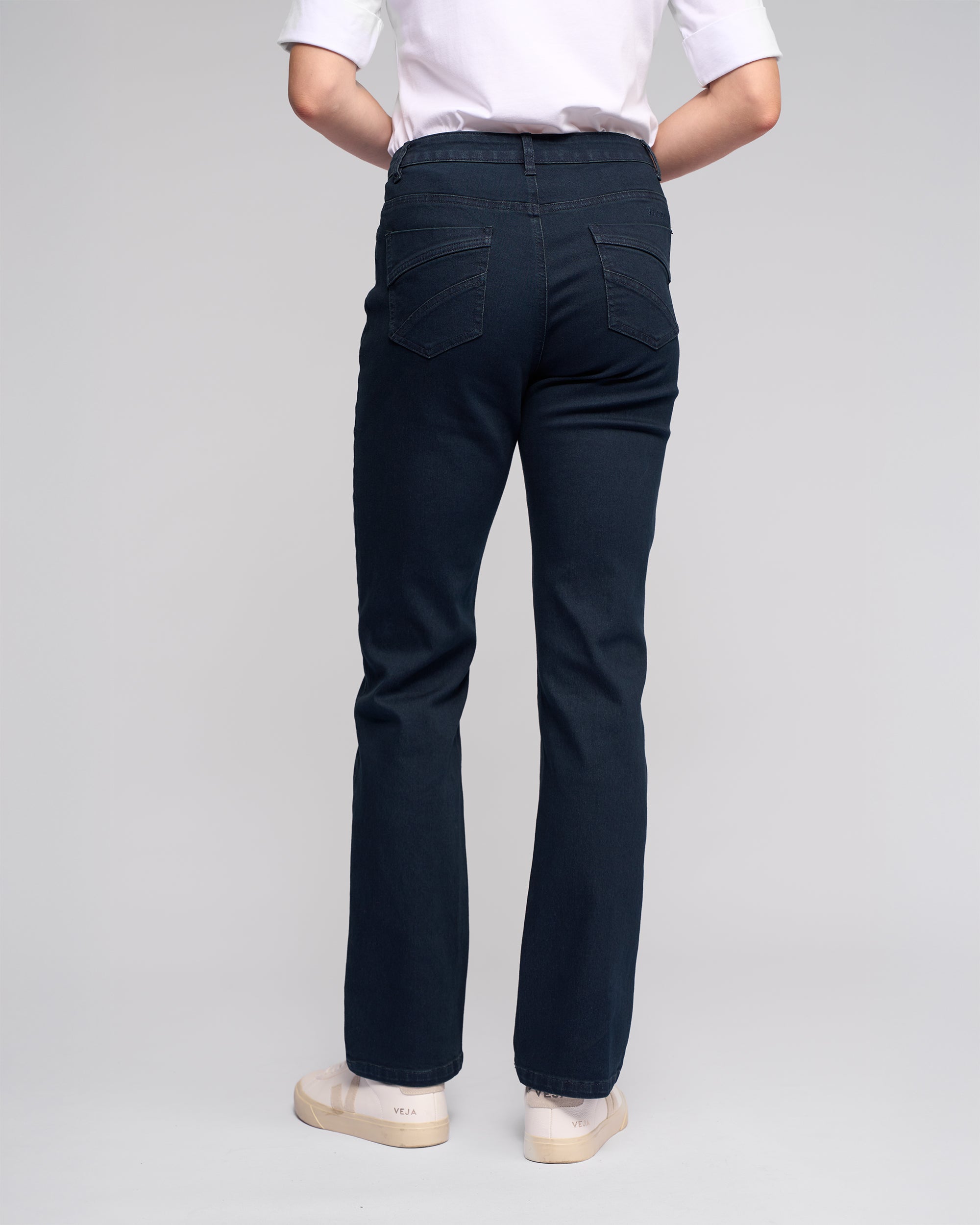 A person stands with their back to the camera, wearing Vassalli’s Radio Jean in indigo denim, a white T-shirt, and light sneakers against a plain light background.