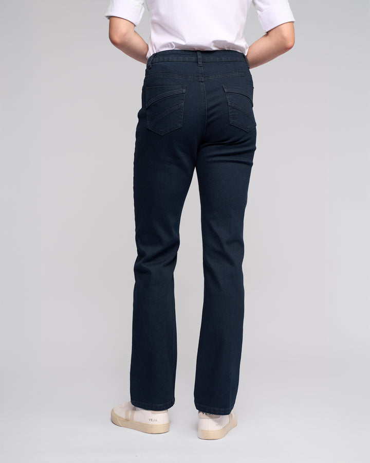 A person stands with their back to the camera, wearing Vassalli’s Radio Jean in indigo denim, a white T-shirt, and light sneakers against a plain light background.