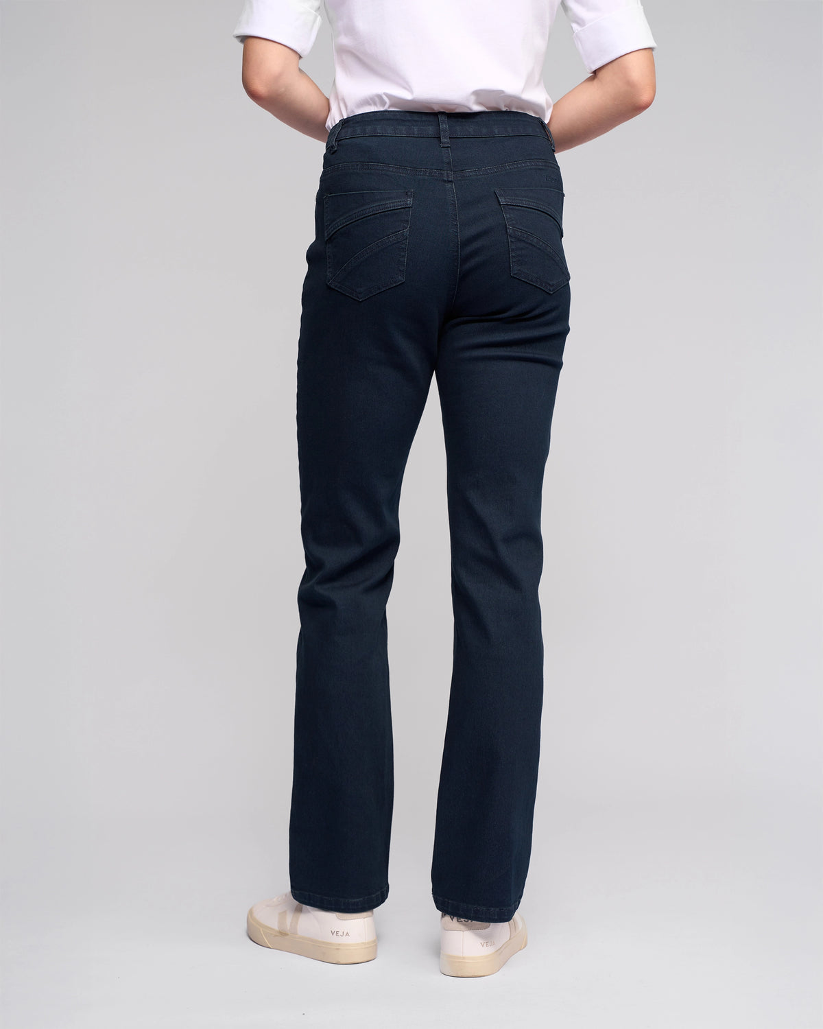 A person stands with their back to the camera, wearing a white shirt, Vassalli Radio Jean in Indigo Denim with a straight-leg fit, and cream-colored sneakers, set against a light gray background.