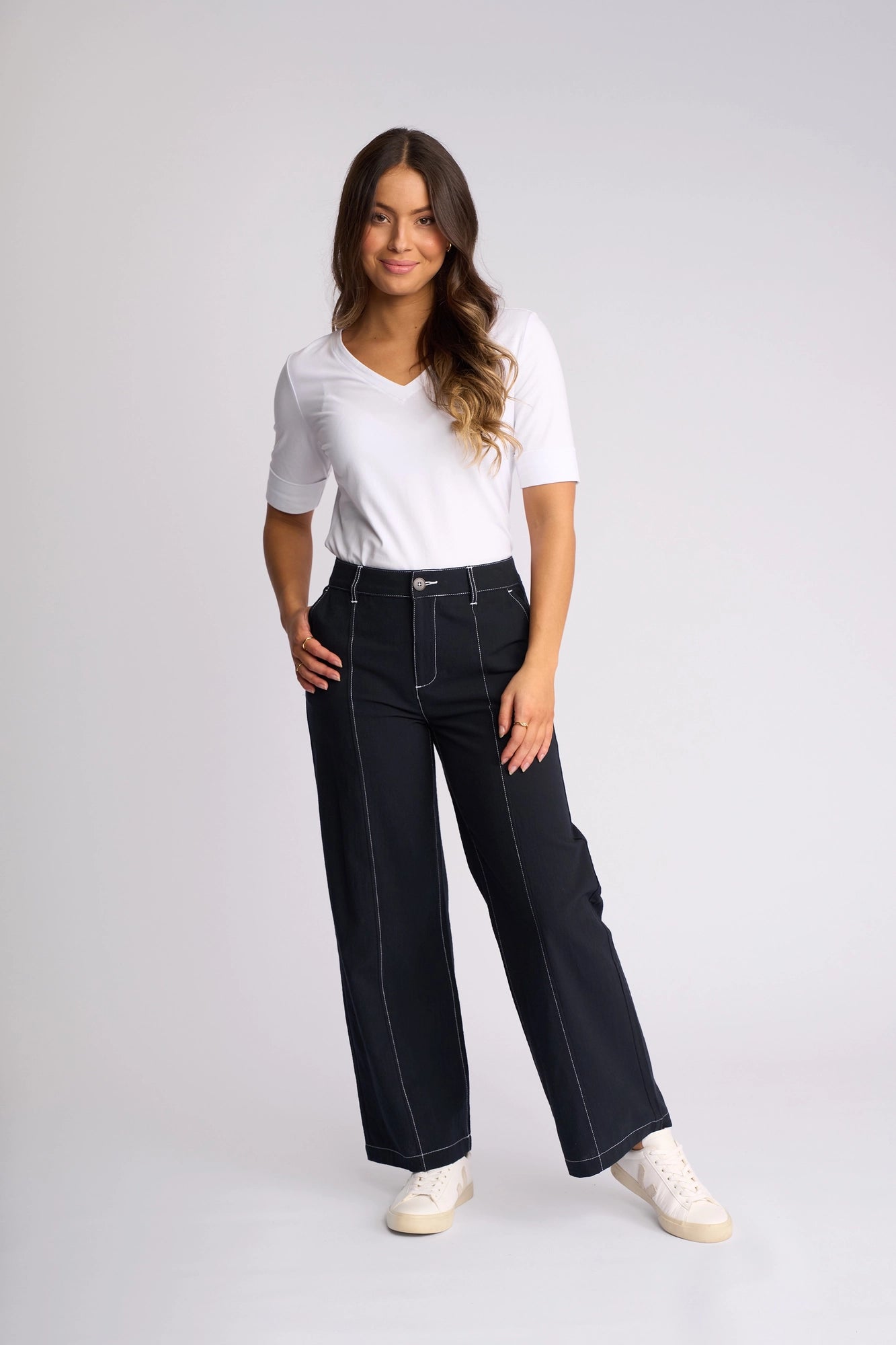 A woman stands smiling, wearing a white short-sleeve top and the Vassalli Oslo Pant in Ink—a high-waisted, 100% cotton, wide-leg ankle-grazer featuring white stitching—paired with white sneakers against a plain light background.