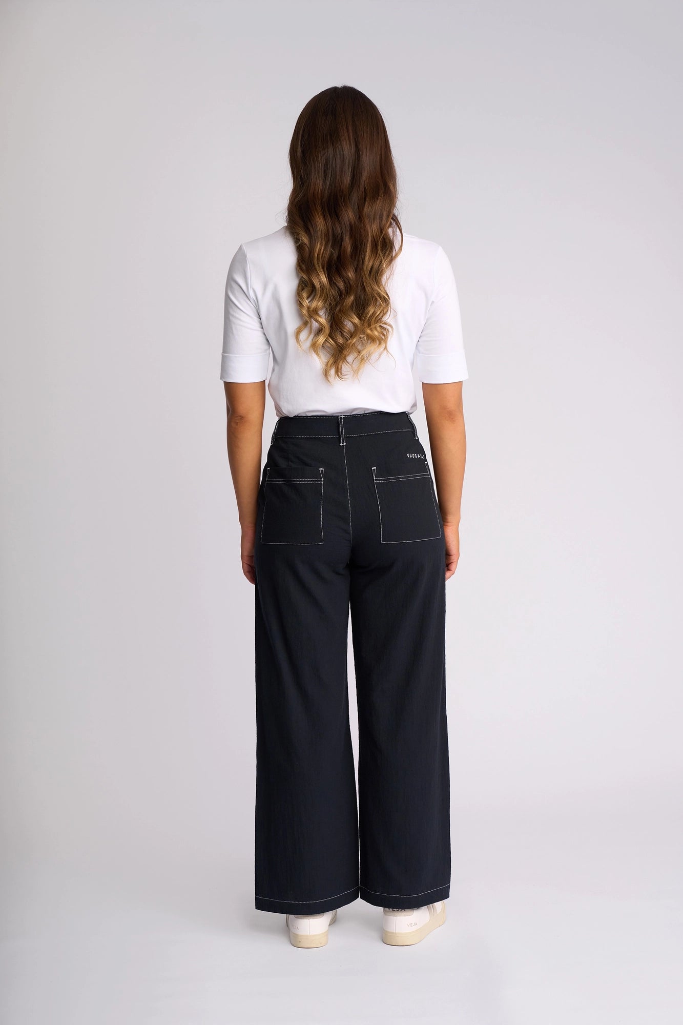 A person with long wavy hair stands facing away, wearing a white shirt and black Vassalli Oslo Pant - Ink—a wide-leg ankle-grazer crafted from 100% cotton with visible stitching—paired with white shoes against a plain light background.