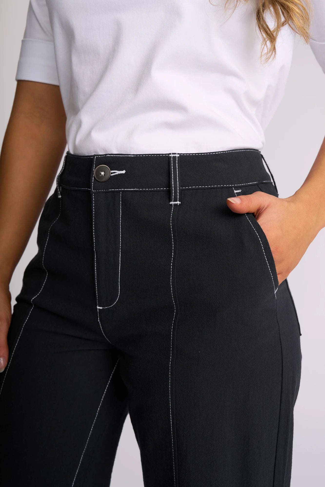 A person wearing a white shirt and black Vassalli Oslo Pant - Ink with white stitching stands with one hand in a pocket. The wide-leg ankle-grazer design, crafted from 100% cotton, adds contemporary flair to the look.