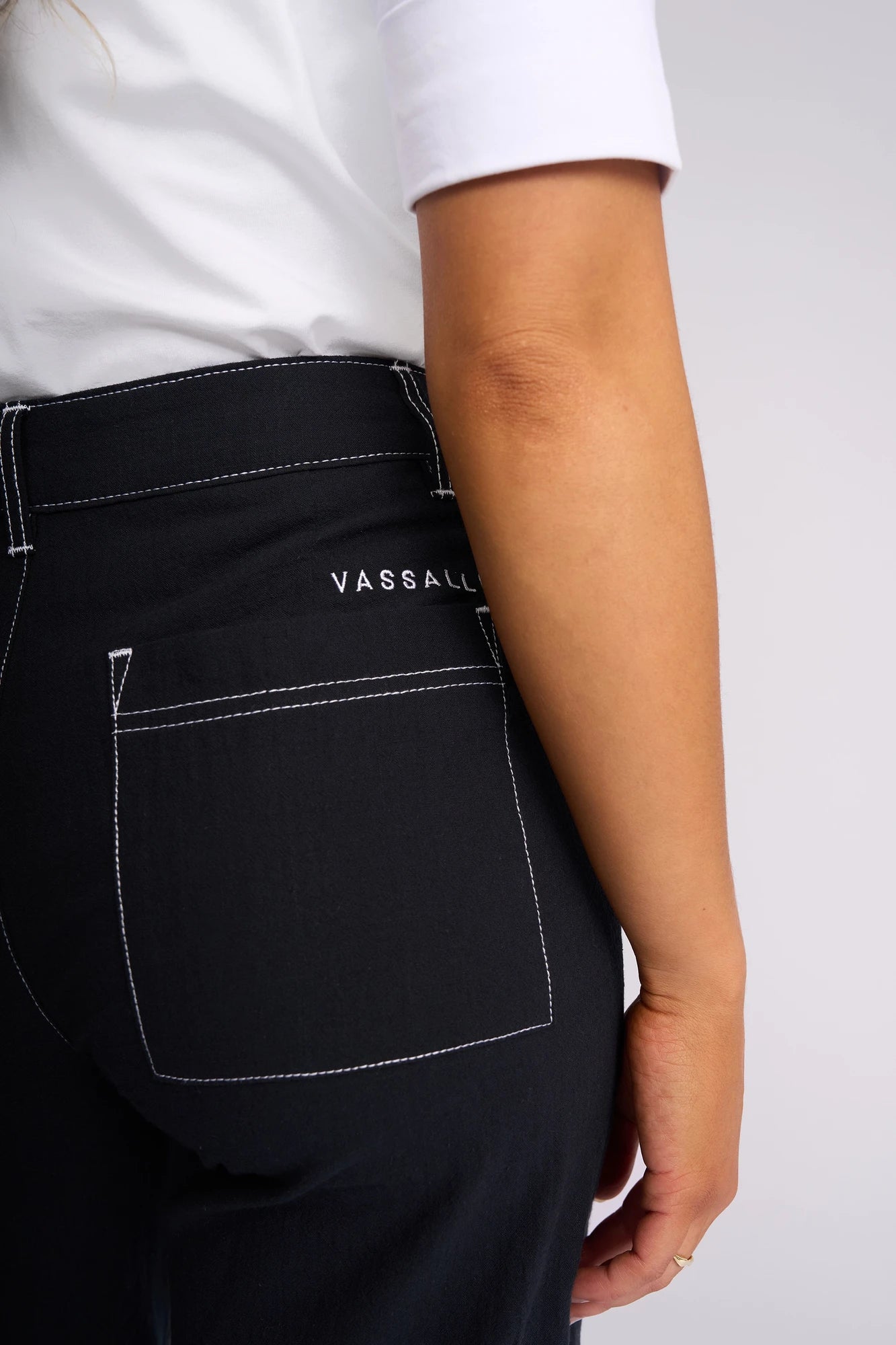 A person wearing a white shirt and black pants with white stitching. The pants, identified as the Vassalli Oslo Pant - Ink, are made from 100% cotton and feature a wide-leg ankle-grazer style with a visible back pocket and the word “VASSALLI” embroidered above it. Only the lower torso and arm are visible.