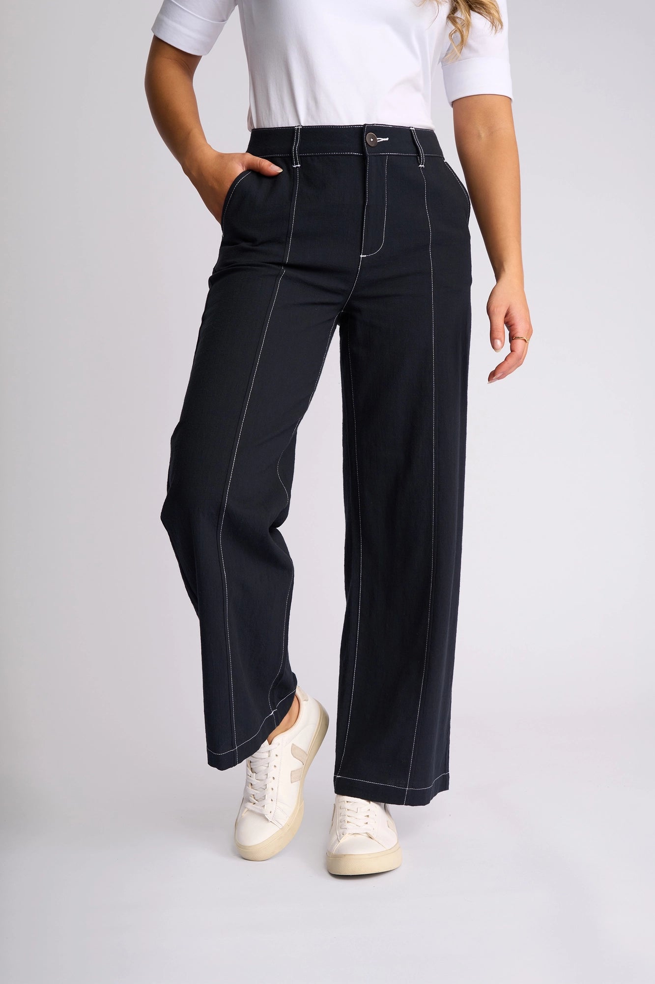 Person wearing a white shirt, Vassalli Oslo Pant in Ink—a wide-leg, ankle-grazer style with white stitching—and white sneakers. Crafted from 100% cotton, the pants feature one hand in a pocket while the other arm rests relaxed at the side. Only the body from shoulder down is visible.