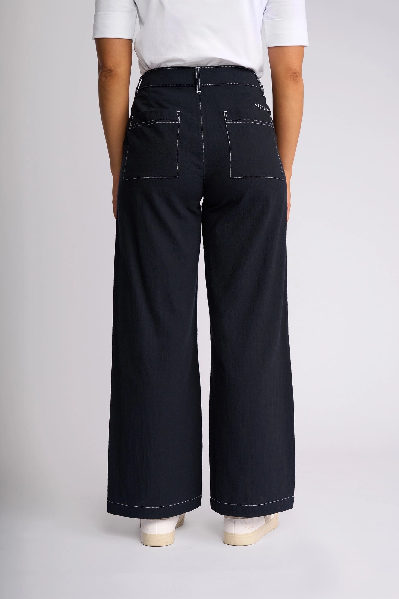 A person stands facing away, wearing a 100% cotton white short-sleeve shirt tucked into high-waisted, wide-leg ankle-grazer Vassalli Oslo Pant in Ink with visible white stitching and patch pockets. White shoes are partially visible. The background is plain and light.