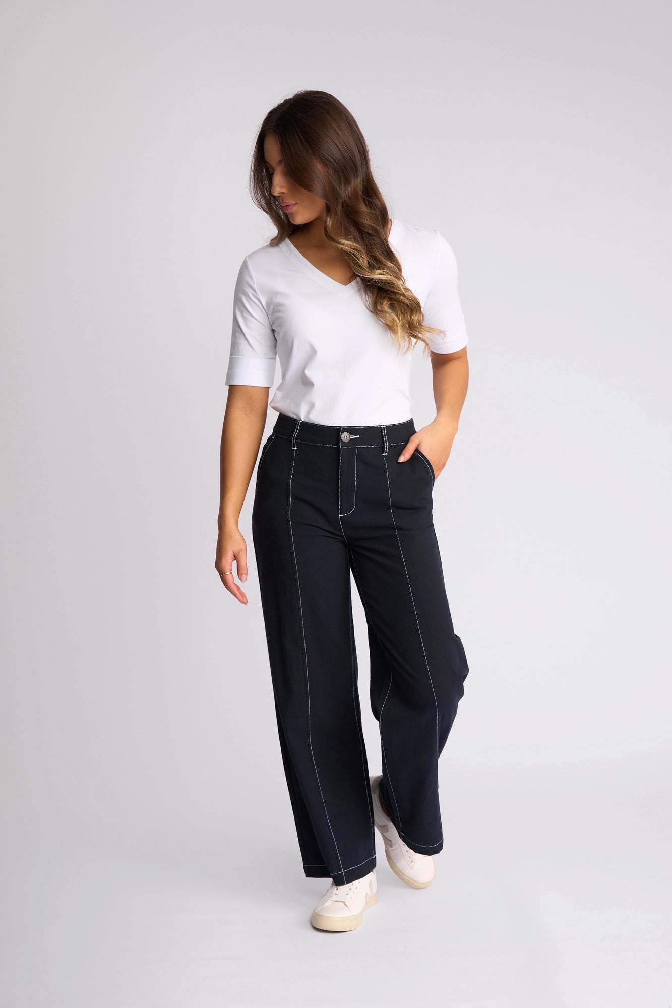 A woman stands against a plain background wearing a white 100% cotton short-sleeve top, the Vassalli Oslo Pant in Ink—a high-waisted, wide-leg ankle-grazer style with distinctive white stitching—and white sneakers. She looks down, with one hand in her pocket and the other by her side.