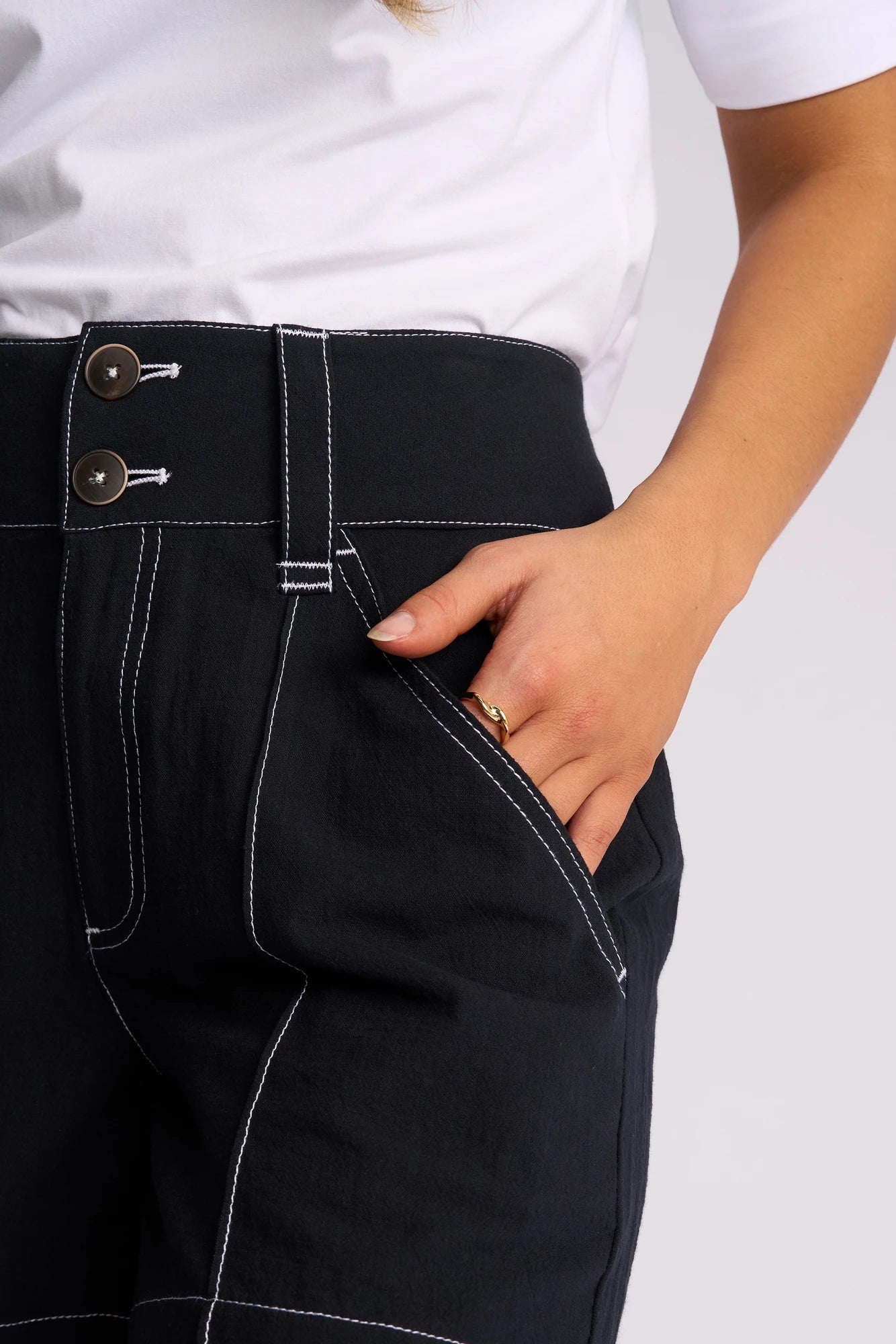 Wearing the Vassalli Lucy Short in Ink, styled effortlessly for summer, this person pairs the black shorts featuring white stitching and two visible waistband buttons with a white shirt. Their right hand is tucked casually into the pocket, displaying a gold ring.