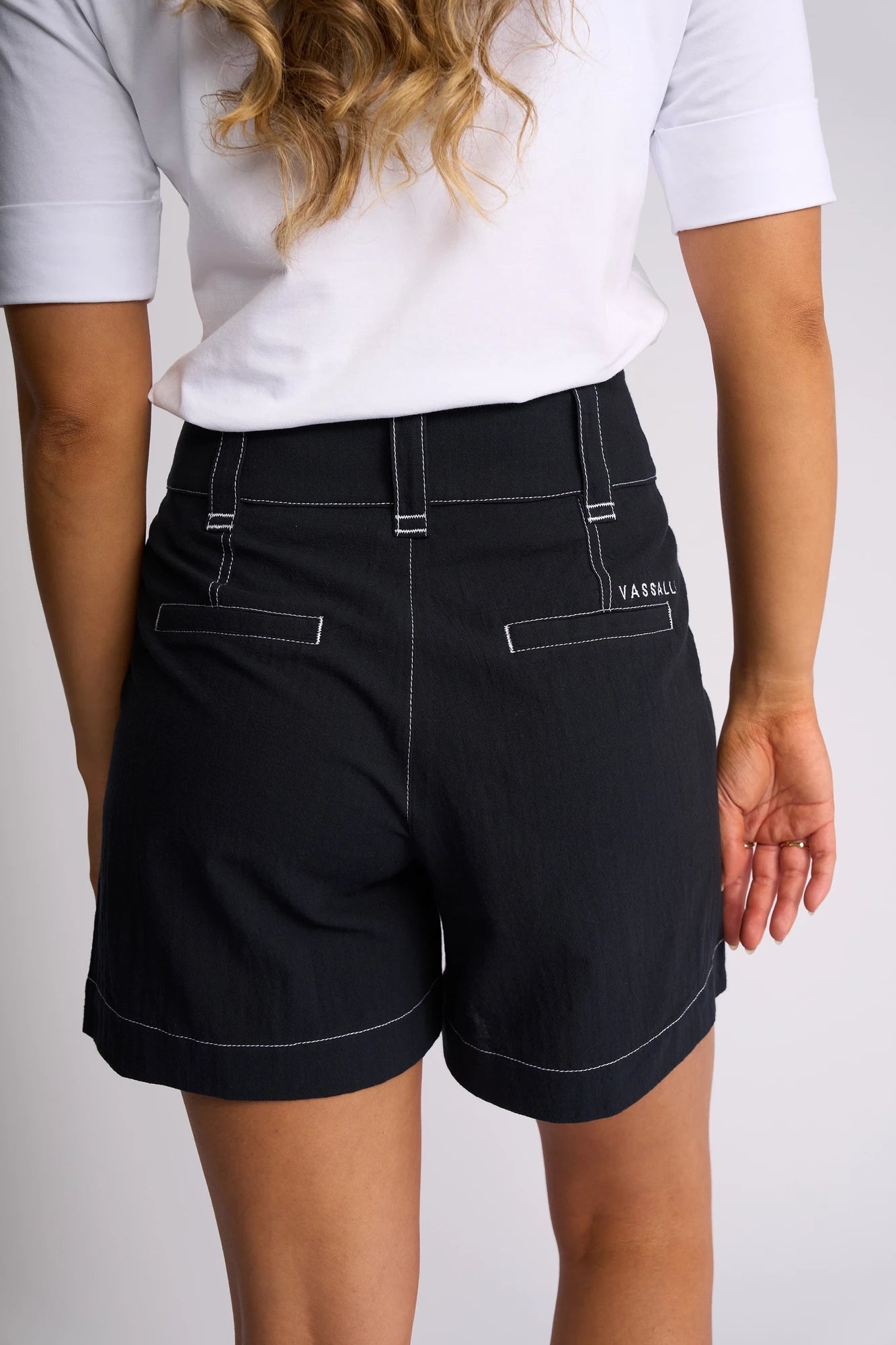A person with wavy blonde hair is seen from behind wearing a white shirt and Vassalli’s Lucy Short - Ink, featuring white stitching. Made from 100% cotton for comfortable summer styling, the shorts offer two back pockets and a small VASSALLI label on the right side.