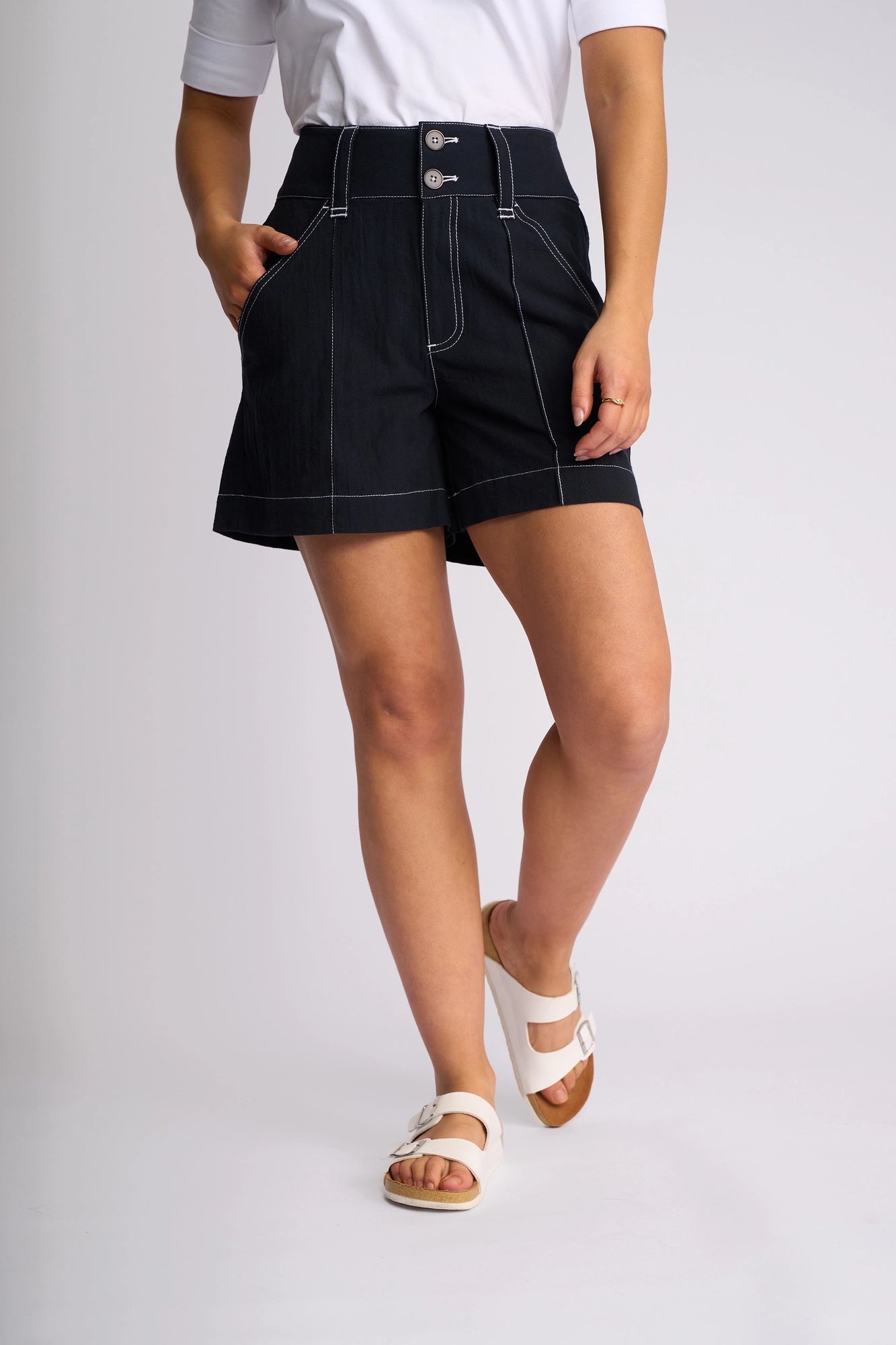 A person wearing a white short-sleeve top, the Vassalli Lucy Short - Ink (a high-waisted black short with white stitching made from 100% cotton), and white sandals stands with one hand in their pocket against a plain background—perfect for effortless summer styling.