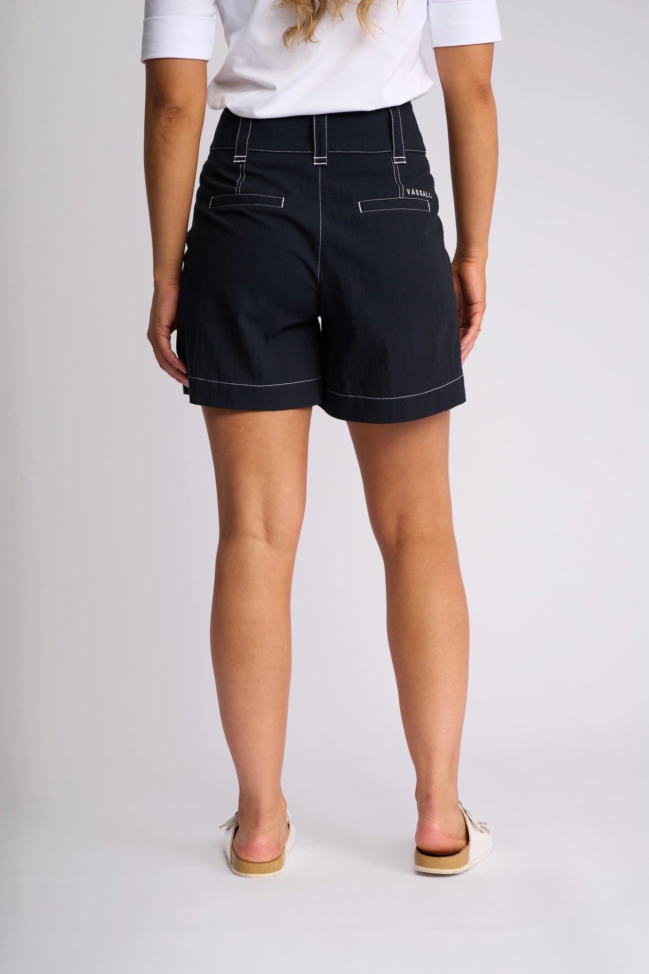 A person stands facing away, wearing a white shirt and the Vassalli Lucy Short - Ink, featuring high-waisted black shorts with white stitching—perfect for effortless summer styling. They are also wearing light-colored slip-on shoes.
