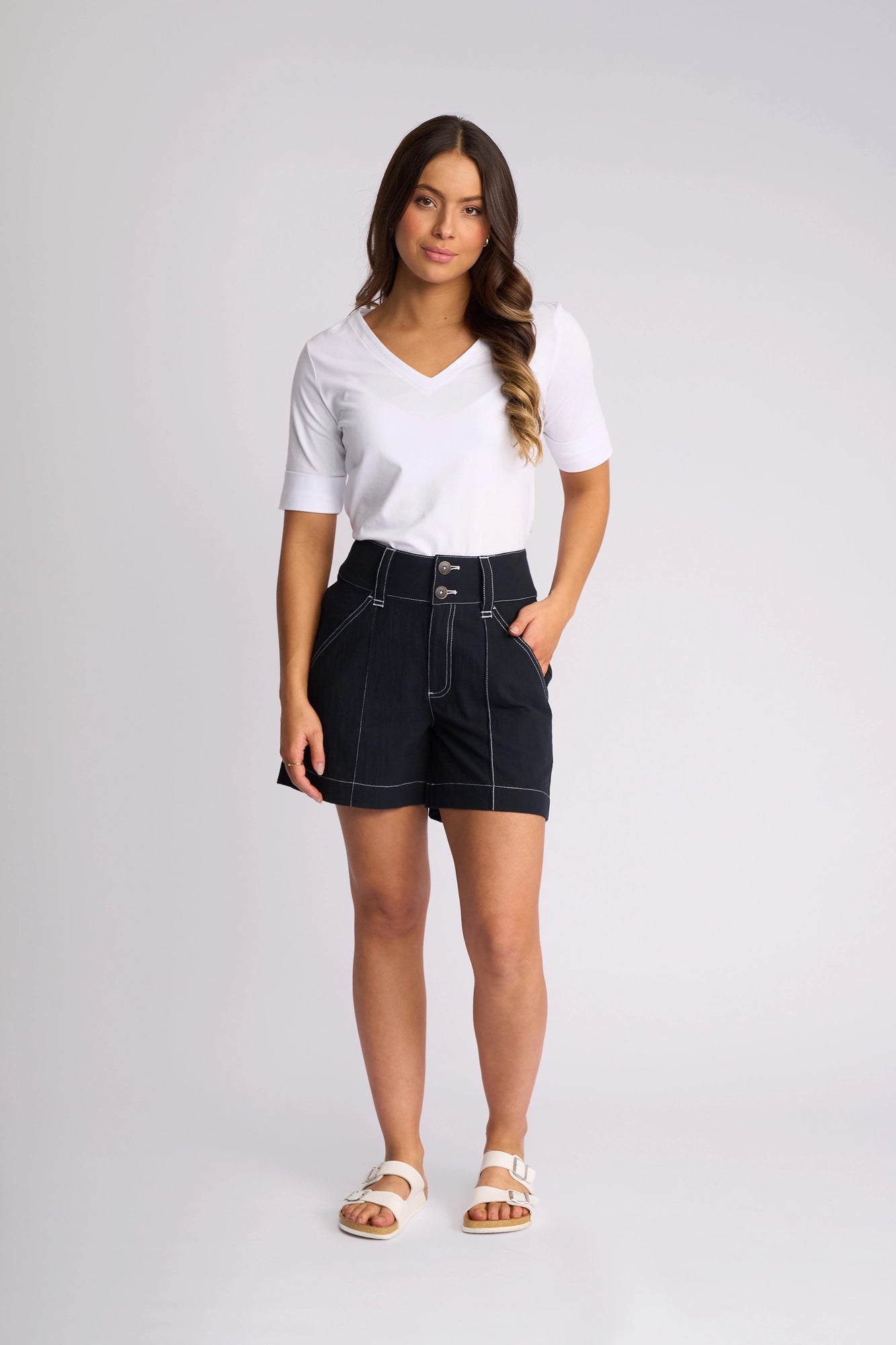 A woman stands against a plain background, wearing a white short-sleeve top, the Vassalli Lucy Short - Ink—high-waisted and crafted from 100% cotton with contrast white stitching—and white sandals. One hand is in her pocket and she faces the camera with a neutral expression, highlighting effortless summer styling.