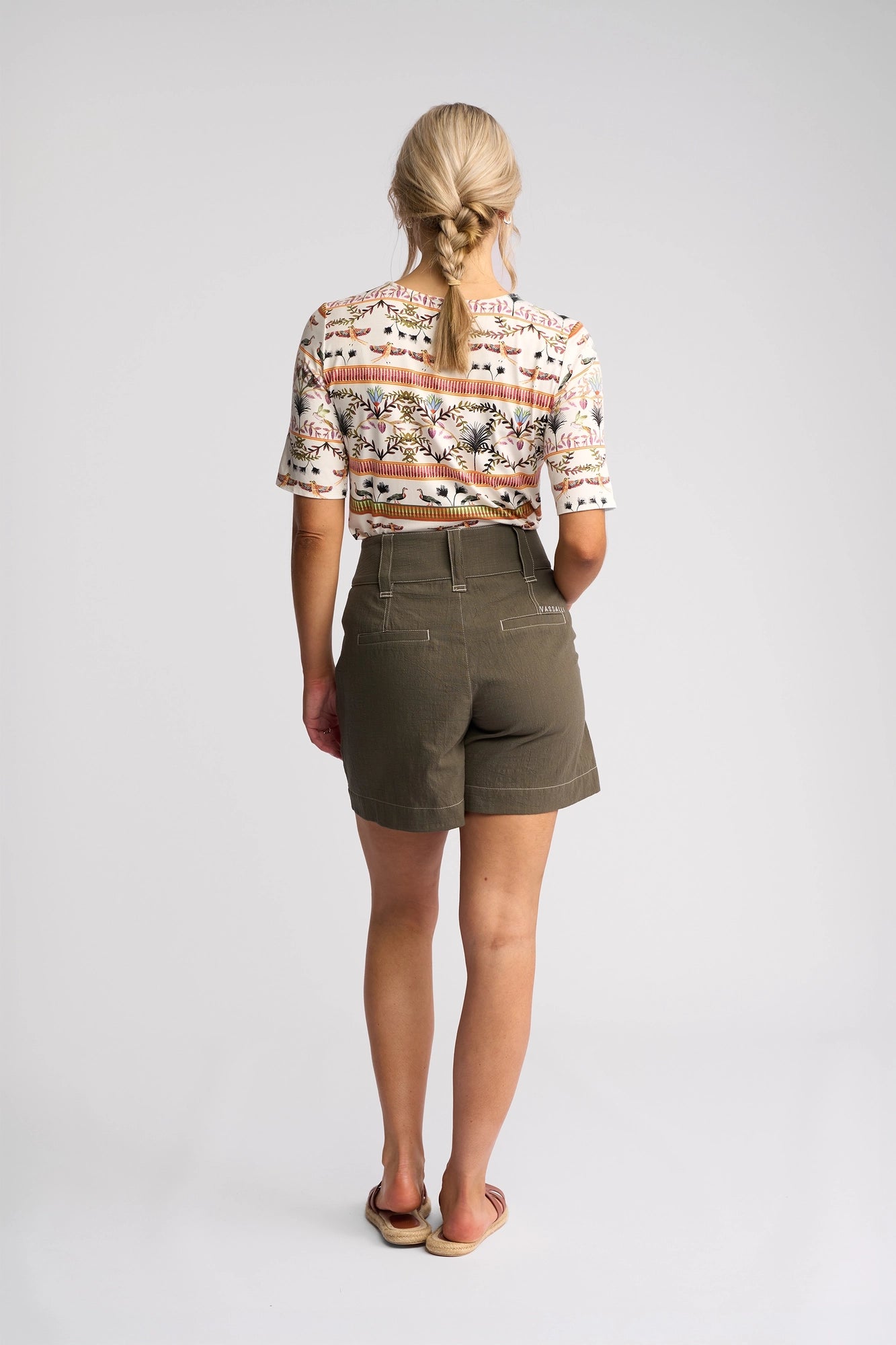 A woman with blonde hair in a low braid, wearing a patterned shirt, the Vassalli Lucy Short in olive, and sandals stands facing away in a studio with a plain white background.