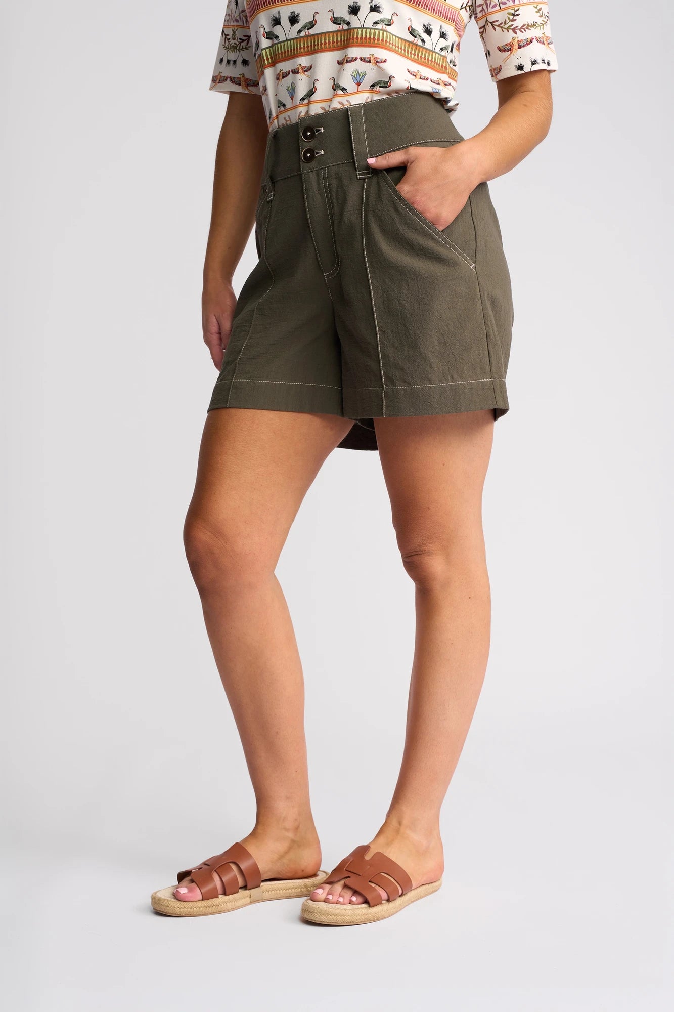 A person stands against a plain light background with one hand in their pocket, wearing the Vassalli Lucy Short in Olive—a wide-leg summer short crafted from linen-look cotton—styled with a patterned short-sleeve top and brown sandals.