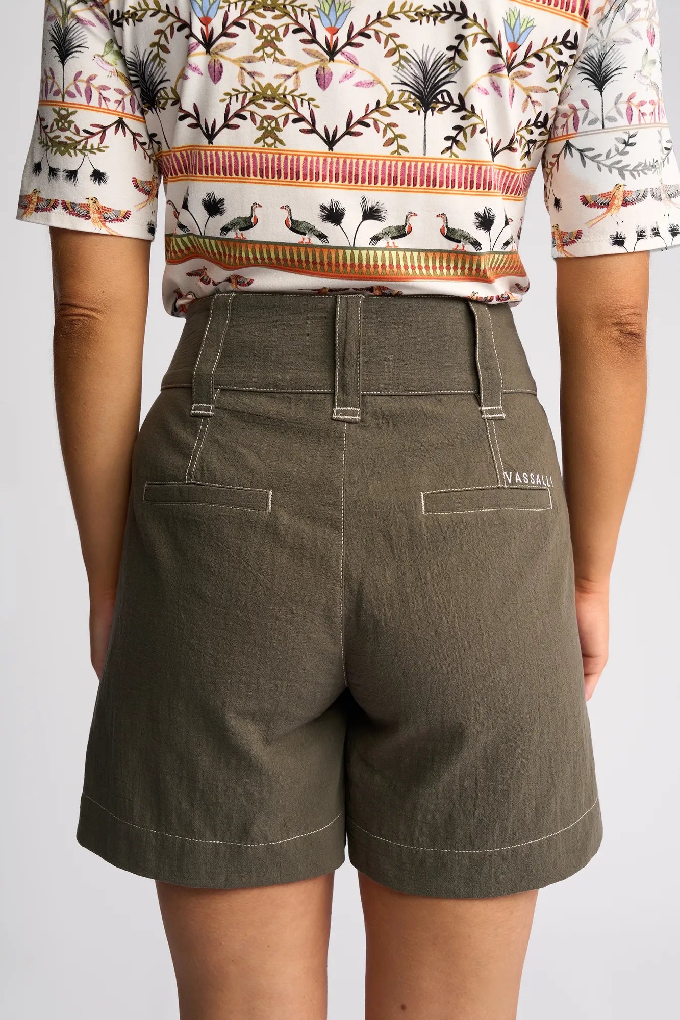 A person is shown from the back wearing the Vassalli Lucy Short in olive, featuring a wide-leg silhouette in linen-look cotton with white stitching, paired with a patterned shirt adorned with birds and plants.