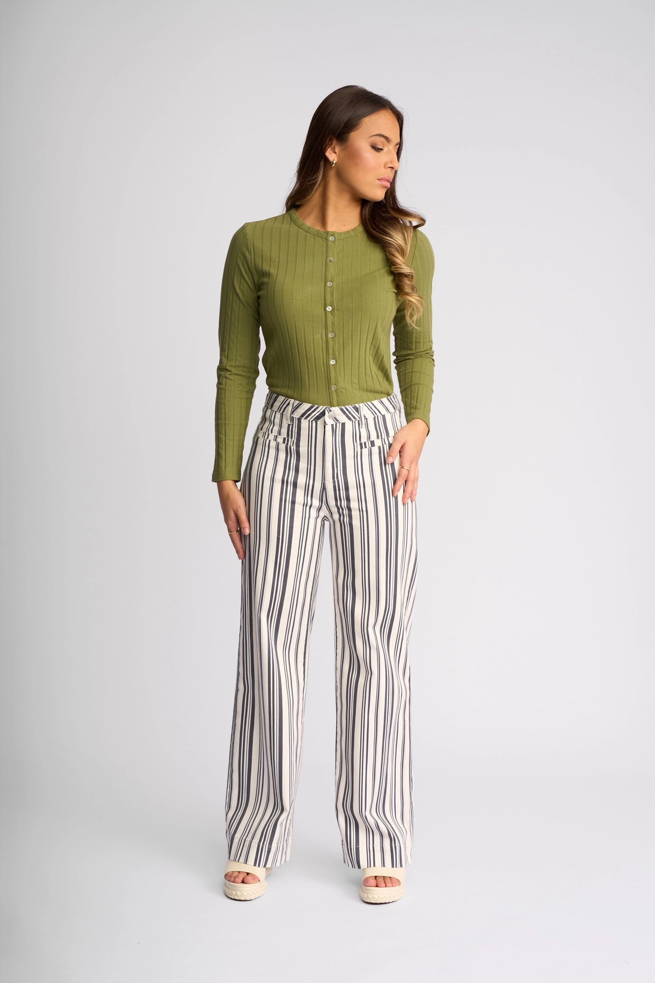 A woman stands against a plain background, wearing a green long-sleeve button-up top paired with Vassalli’s Rune Jean in Multi Stripe Denim—high-waisted, wide-leg pants featuring vertical black and white stripes. She looks to her left with her hands at her sides, embodying effortless summer style.