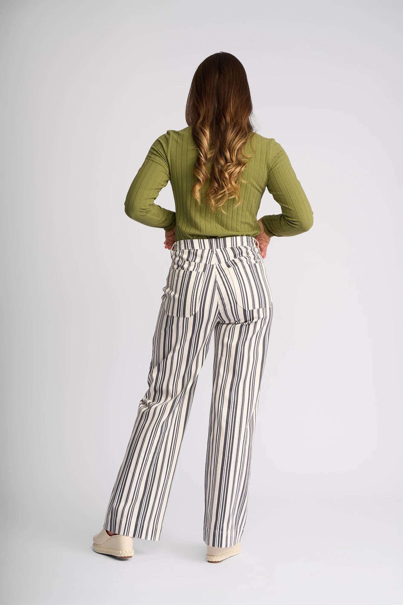 A person stands facing away, wearing a green long-sleeve top paired with Vassalli’s Rune Jean in Multi Stripe Denim—high-waisted, wide-leg jeans featuring vertical black and white stripes. With long, wavy brown hair and light-colored shoes, they perfectly showcase a summer denim look against a plain background.