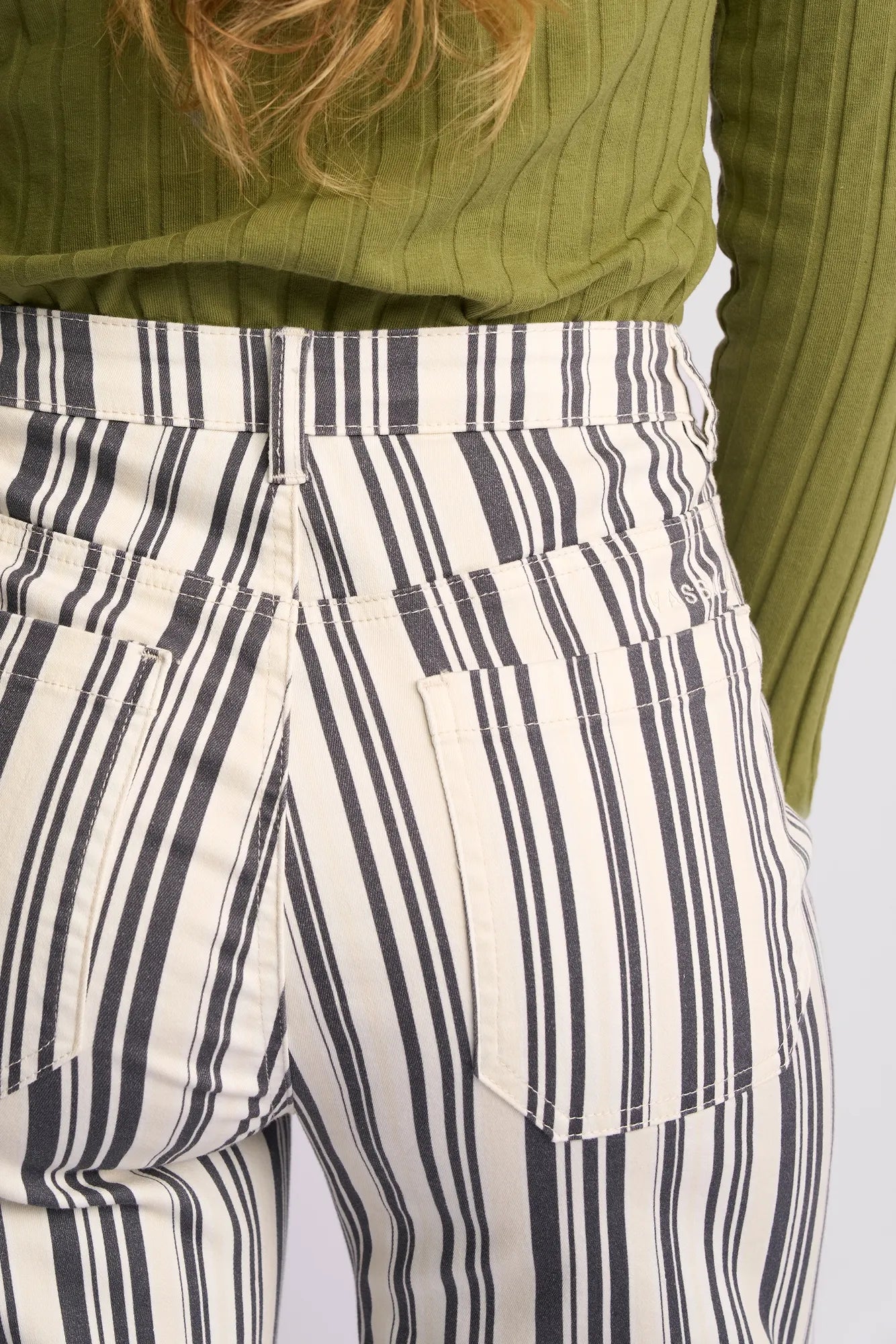 A person shown from the back is wearing Vassalli’s Rune Jean in Multi Stripe Denim—featuring black and white vertical stripes—paired with a green ribbed long-sleeve shirt.