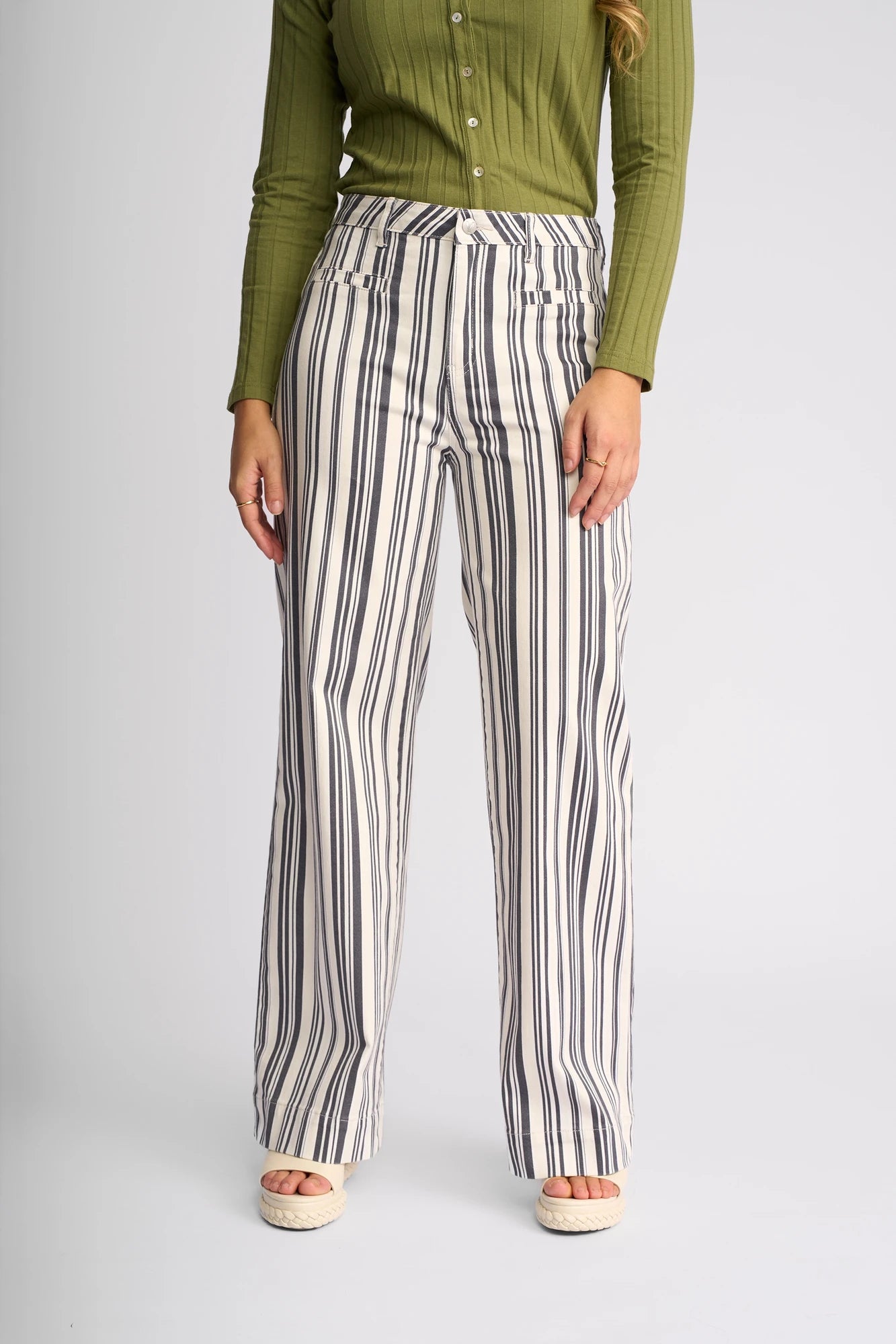 A person wearing a long-sleeve, ribbed, olive-green top and the Vassalli Rune Jean in Multi Stripe Denim—a high-waisted, wide-leg pant featuring vertical black and white stripes—stands against a plain light background.