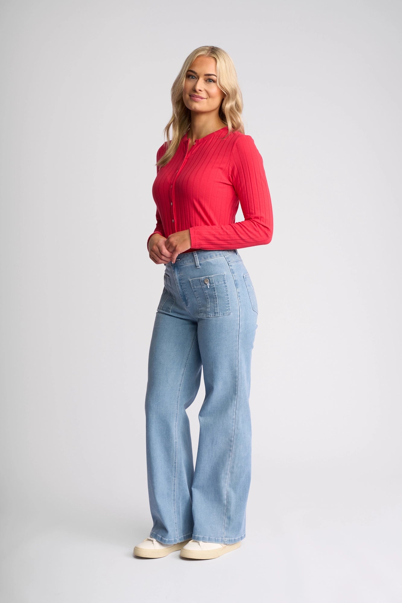 A woman with long blonde hair wears a red long-sleeve top, Vassalli’s Lottie Jean in light wash denim, and white shoes, standing against a plain light gray background and smiling softly with her hands loosely clasped.