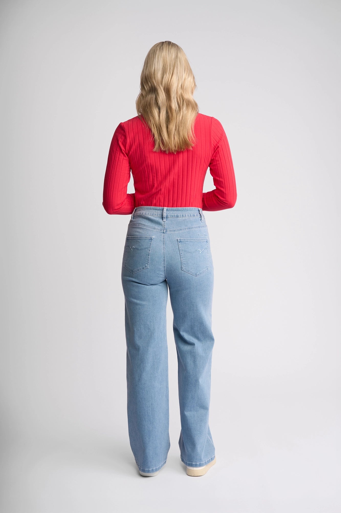 A woman with long blonde hair stands facing away, wearing a red long-sleeve top and the Vassalli Lottie Jean in light wash denim, featuring a high-rise fit and back pockets. The background is plain and light-colored.