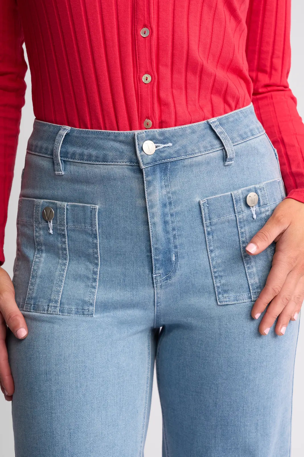A person wearing the Vassalli Lottie Jean in light wash denim, featuring a high-rise, wide-leg fit with large front pockets, paired with a red ribbed button-up top. Only the torso and upper legs are visible.