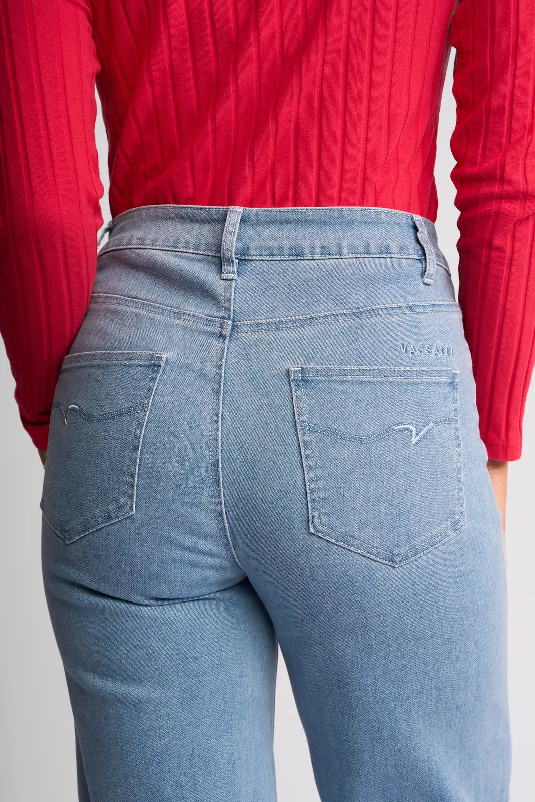 A person is seen from the back wearing Vassalli’s Lottie Jean in light wash denim, featuring wide legs, two back pockets, and a subtle embroidered design on the right pocket, paired with a red ribbed long-sleeve top.