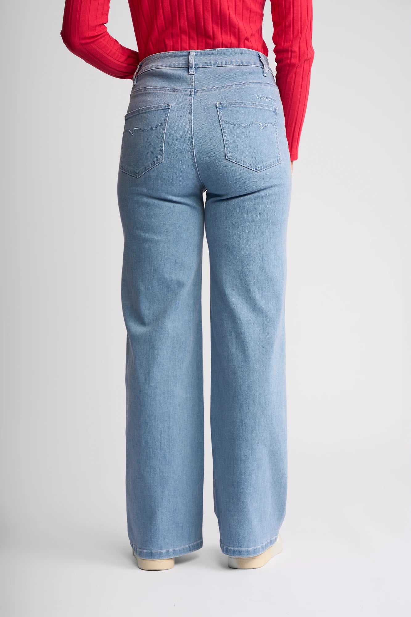 A person is wearing the Vassalli Lottie Jean - Light Wash Denim, which features a high-waisted wide-leg silhouette and a high-rise fit, paired with a red ribbed long-sleeve top, shown from the back against a plain white background.