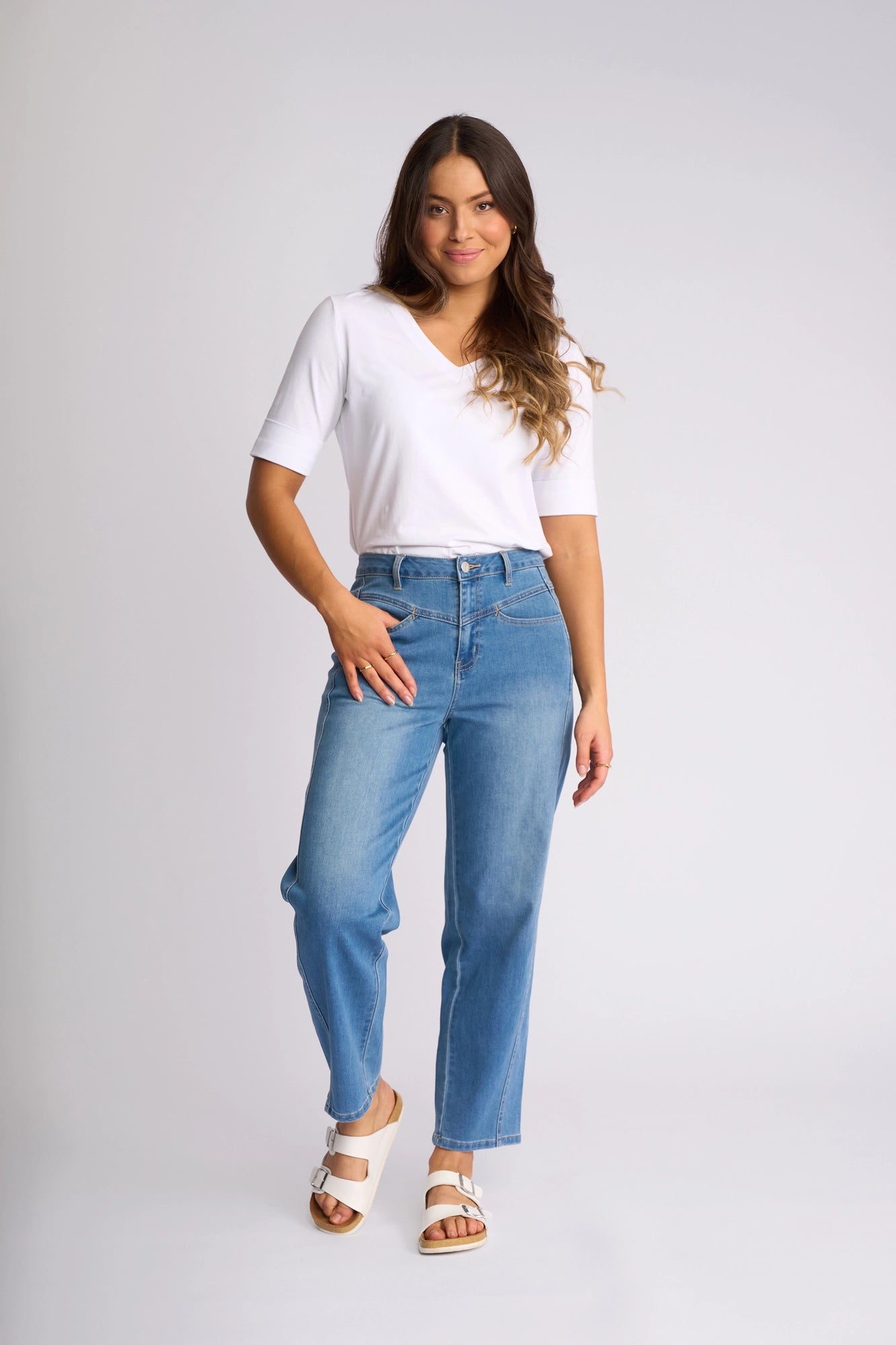 A woman with long brown hair, smiling with one hand in her pocket, wears a white short-sleeve top, white sandals, and the Vassalli Apollo Jean - Vintage Denim, standing against a plain light background.