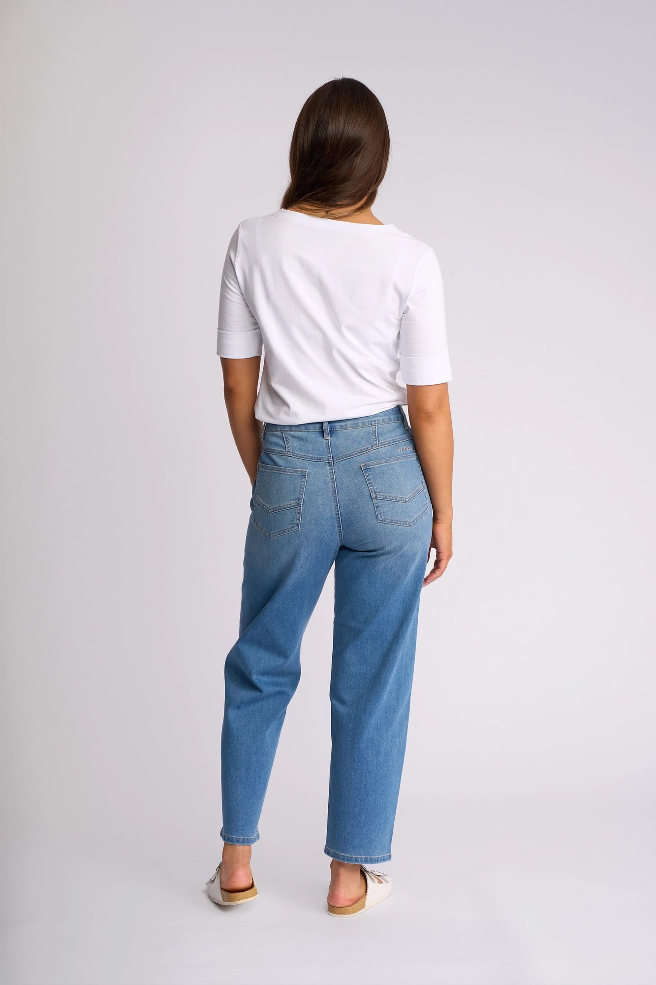 A woman stands facing away against a plain light background, wearing a white short-sleeve shirt, white shoes, and Vassalli’s Apollo Jean in vintage denim—a light blue, high-waisted style.