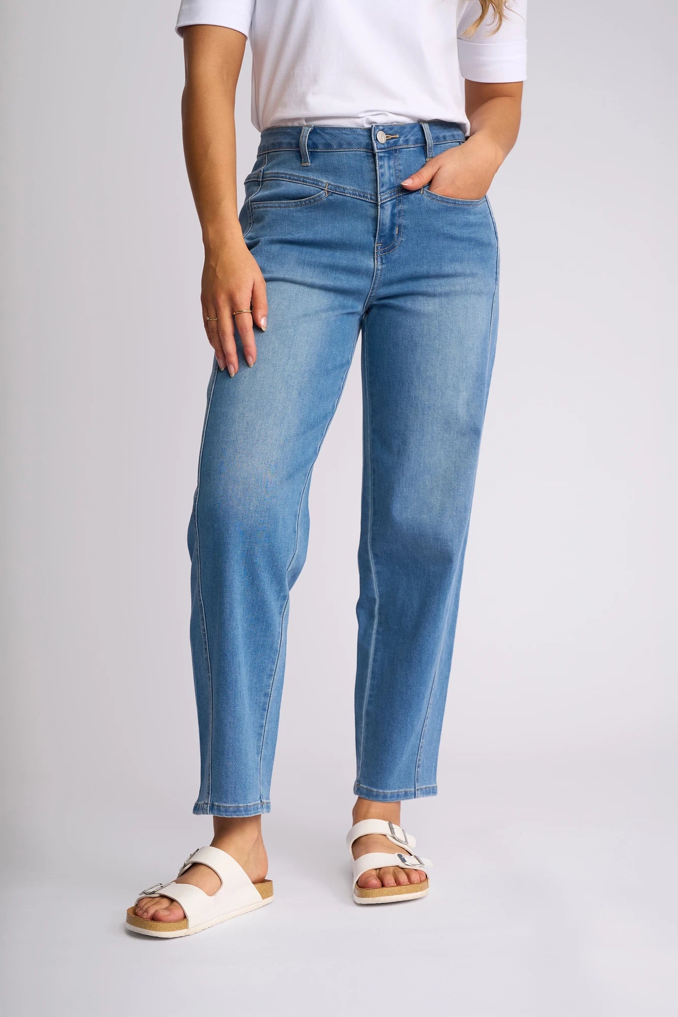 A person wearing a white shirt, Vassalli Apollo Jean - Vintage Denim high-waisted barrel-leg jeans, and white sandals stands against a plain background with one hand in their pocket.