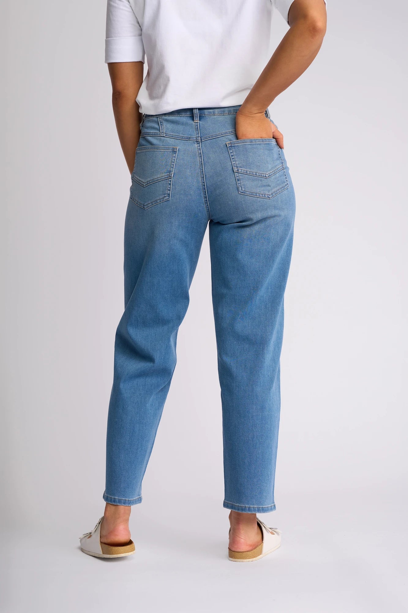 A person wearing Vassalli’s Apollo Jean in Vintage Denim and a white shirt stands with their back to the camera, one hand tucked in a back pocket. They complete the look with white shoes.