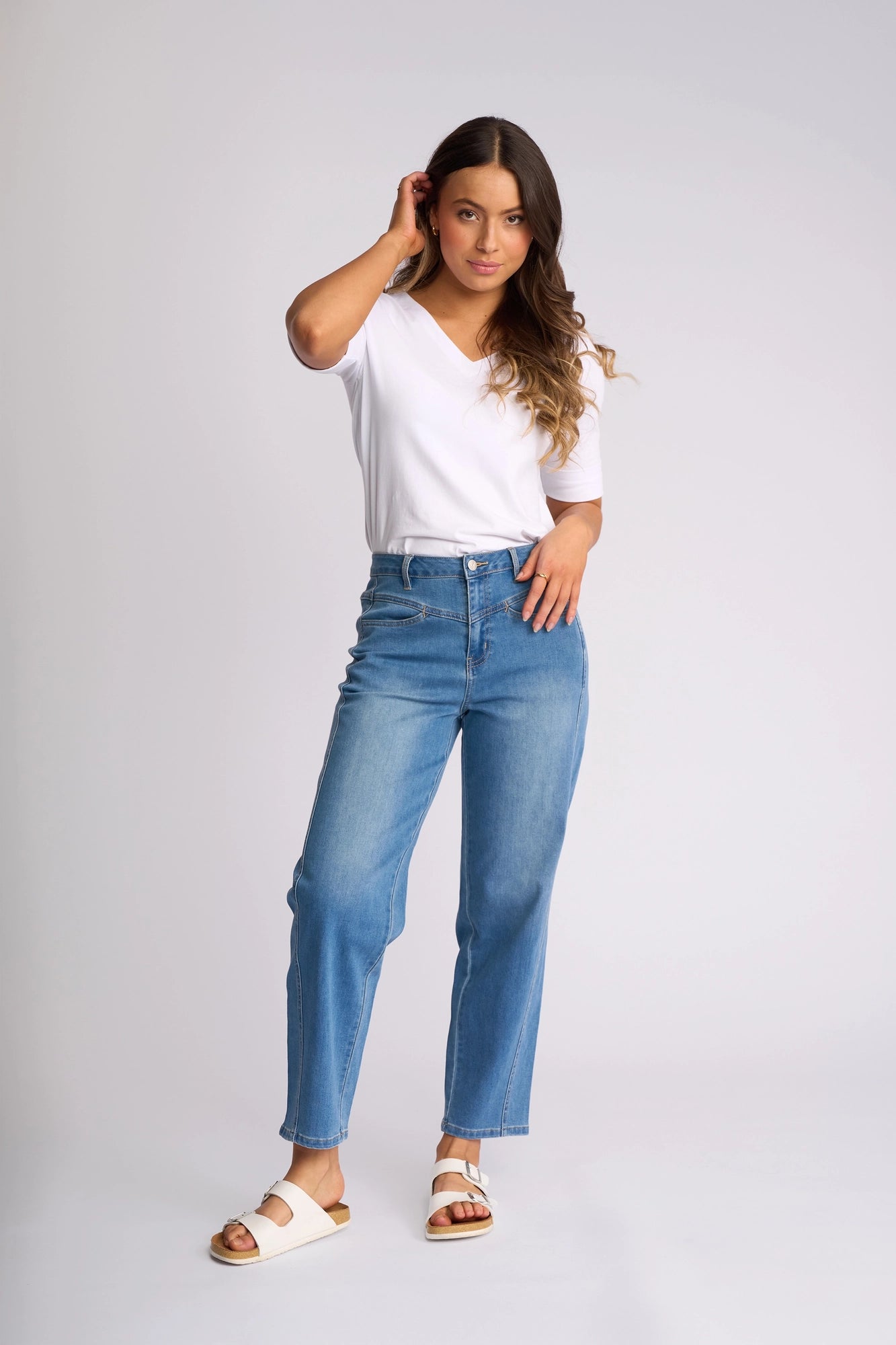 A woman stands against a plain light background, touching her hair with one hand and looking at the camera, wearing a white t-shirt, Vassalli Apollo Jean in Vintage Denim, and white sandals.