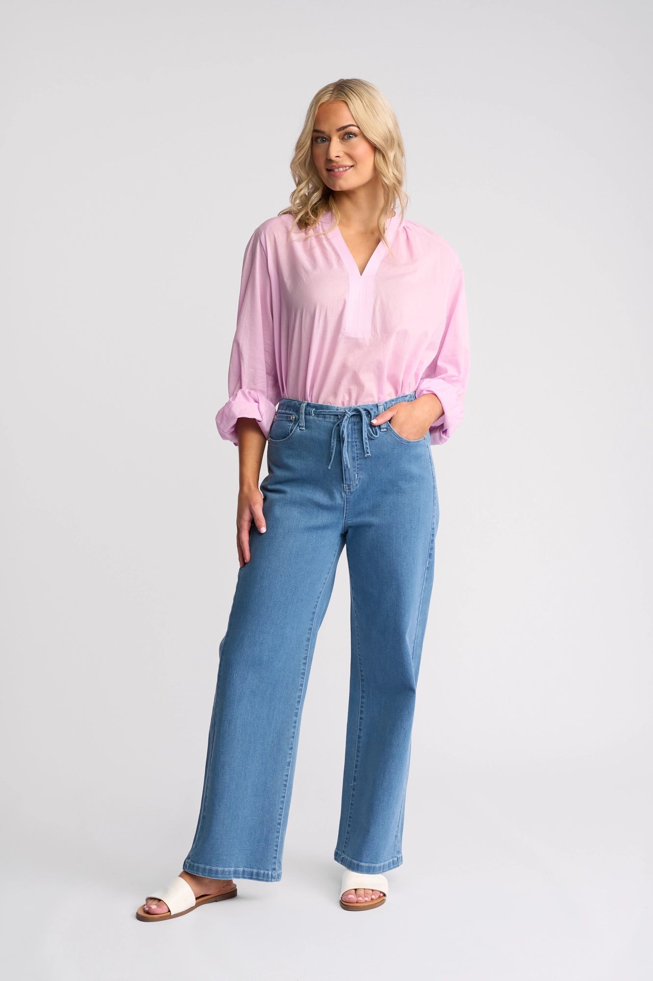 A woman stands in a relaxed pose against a plain light background, wearing a light pink long-sleeve blouse, the Vassalli Yarra Jean - Mid Denim high-waisted wide-leg jeans with an adjustable drawstring waist, and white slide sandals.