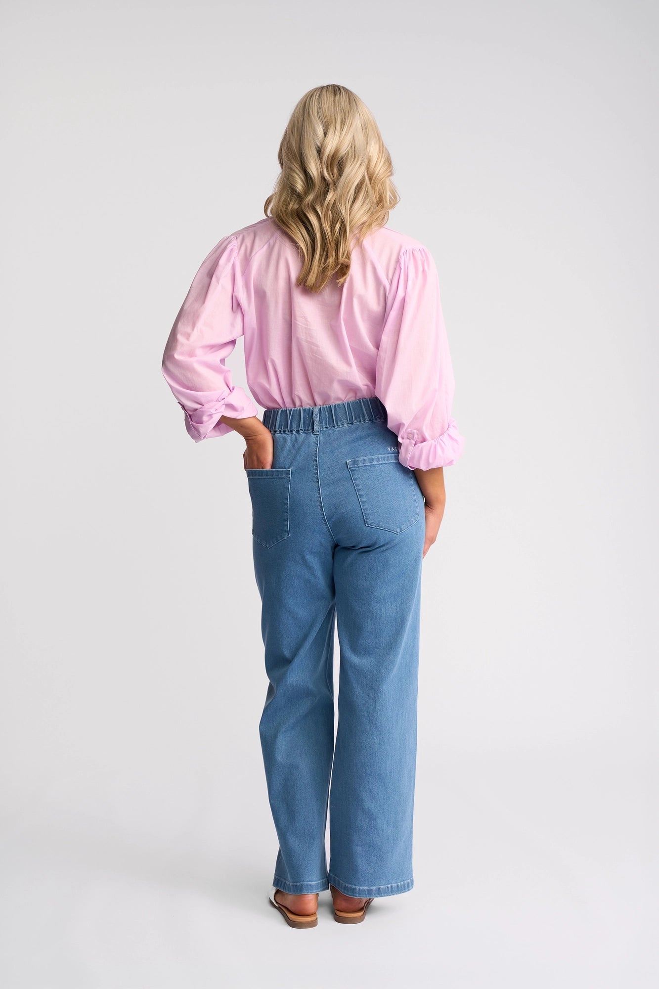 A person with wavy blonde hair stands facing away, wearing a light pink long-sleeve shirt, brown shoes, and the Vassalli Yarra Jean - Mid Denim—high-waisted wide-leg jeans in stretch denim—against a plain light background.