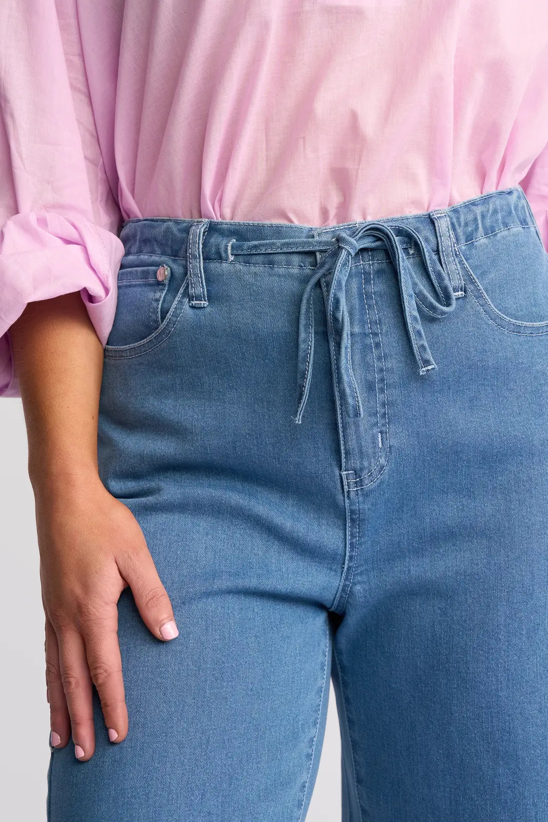 A person is shown from mid-torso to upper thighs wearing the Vassalli Yarra Jean - Mid Denim, featuring light blue stretch denim with an adjustable drawstring waist, paired with a pink long-sleeve shirt with rolled-up sleeves. Their left hand rests beside their hip.