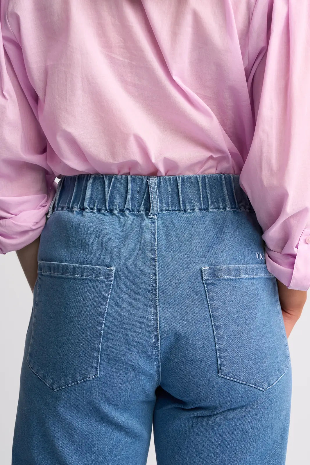 A person wearing a light pink shirt and Vassalli Yarra Jean - Mid Denim with an adjustable drawstring waist, shown from the back.