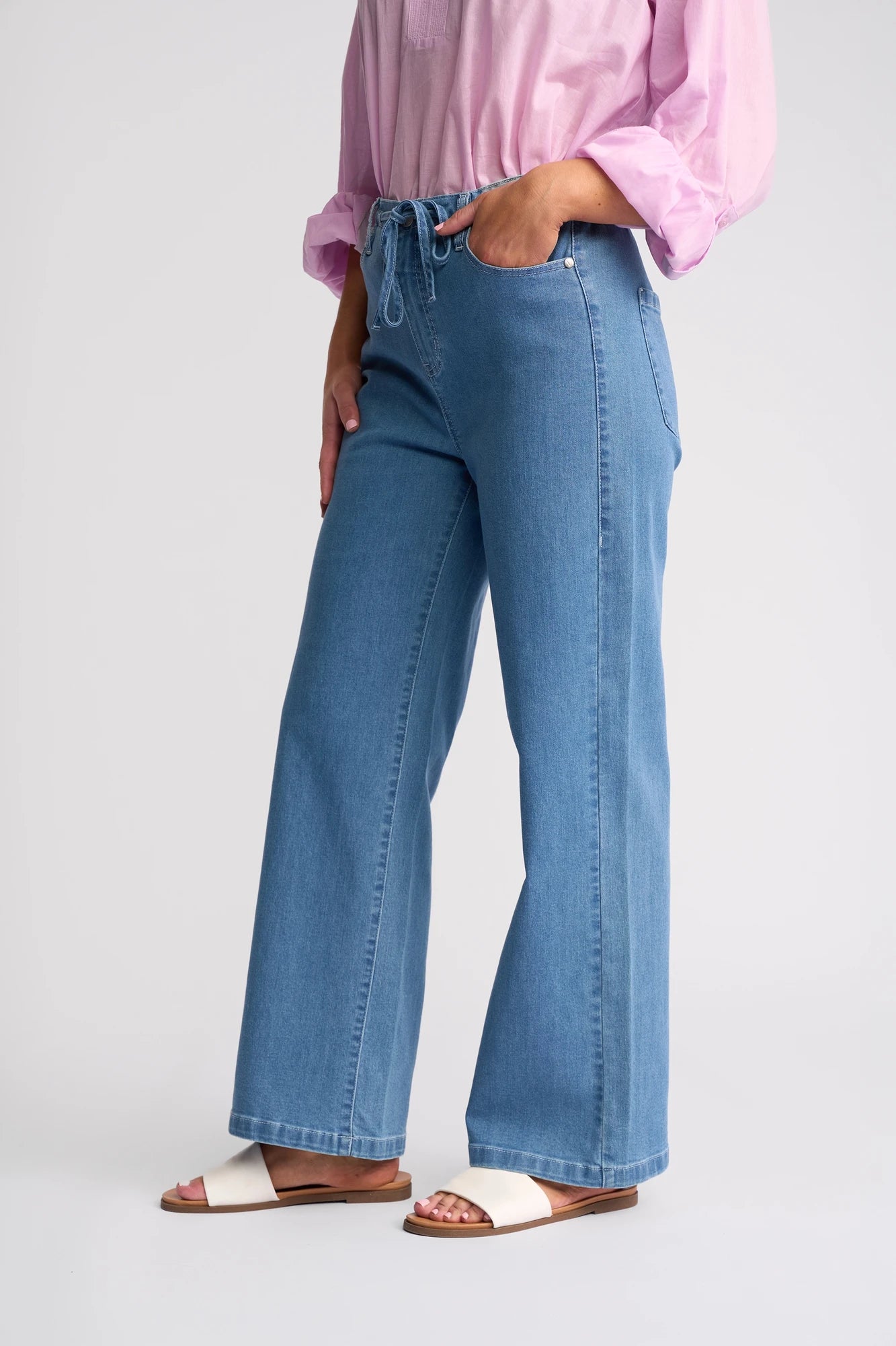 Wearing the Vassalli Yarra Jean - Mid Denim, which features light blue stretch denim and a wide-leg silhouette with an adjustable drawstring waist, this person pairs the jeans with a pink long-sleeve shirt and white sandals, standing casually with one hand in their pocket against a plain background.