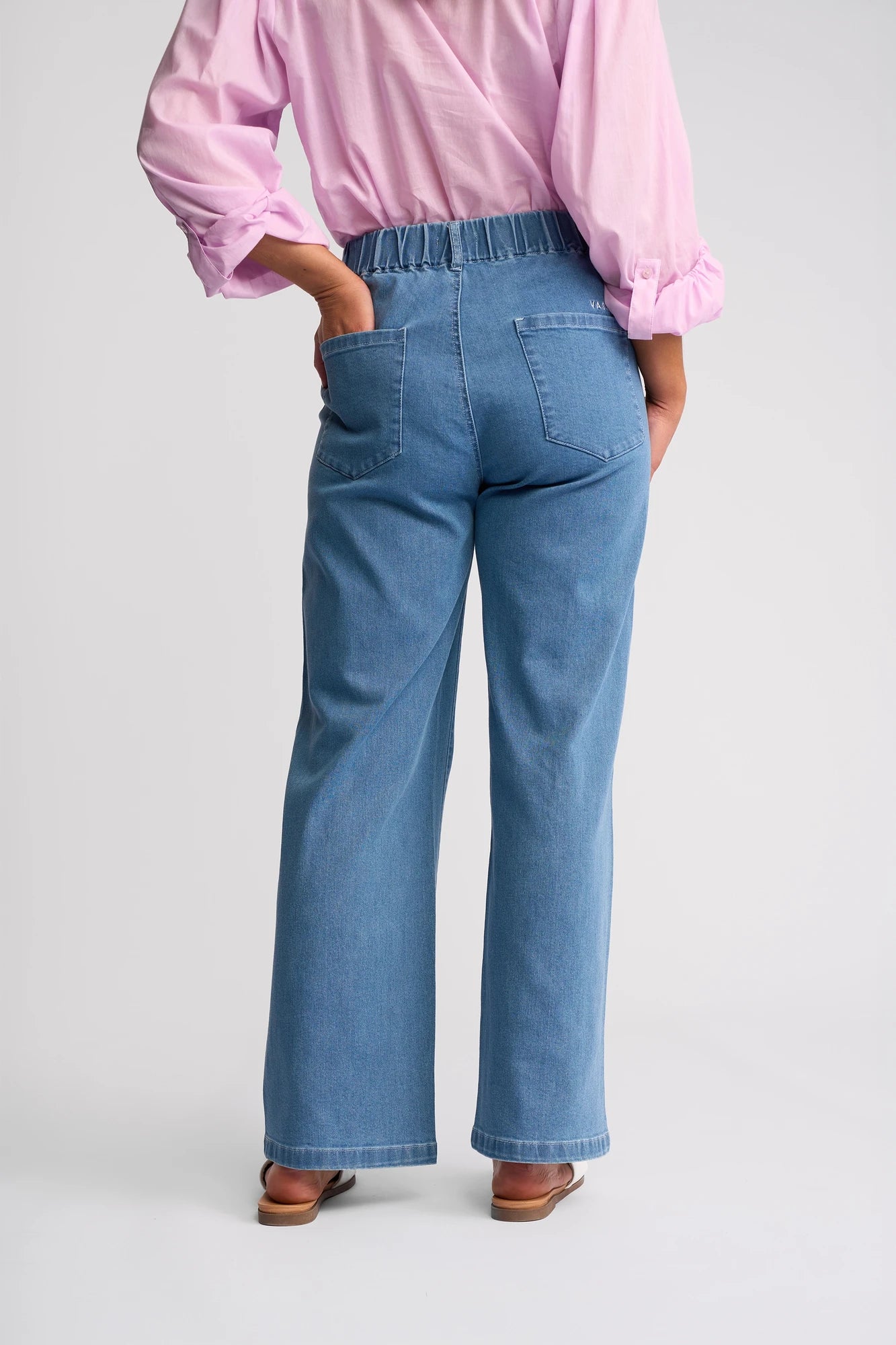 A person is shown from the back wearing Vassalli’s Yarra Jean - Mid Denim, a pair of blue high-waisted wide-leg jeans with an elastic waistband, paired with a light pink long-sleeve shirt and brown sandals. The person stands with one hand on their hip.