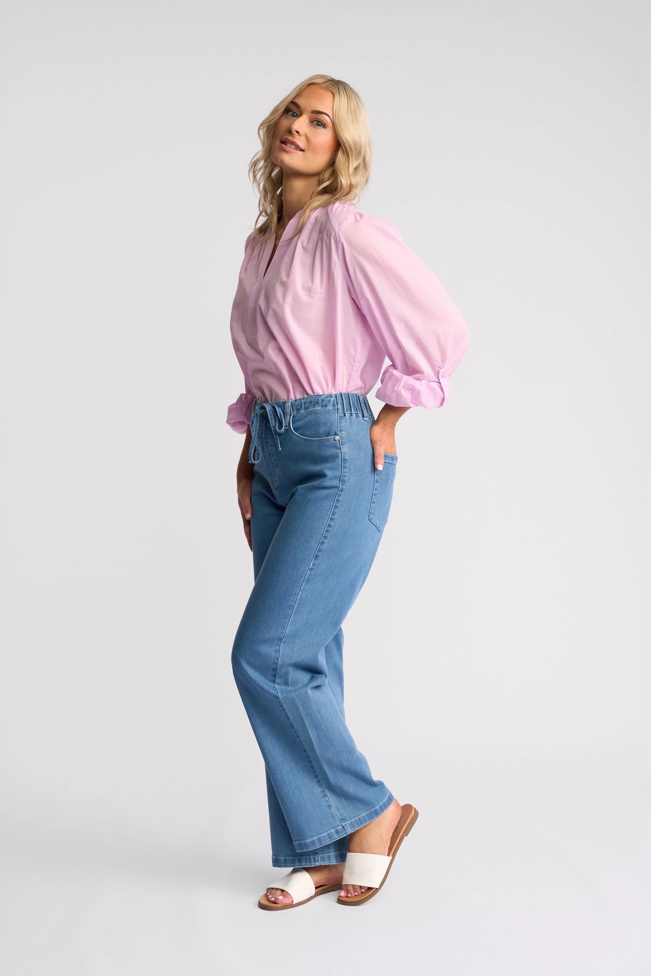 A woman in a pink blouse, wearing Vassalli’s Yarra Jean - Mid Denim—high-waisted, wide-leg jeans with an adjustable drawstring waist—and white sandals, stands sideways with one leg bent and smiles against a plain white background.