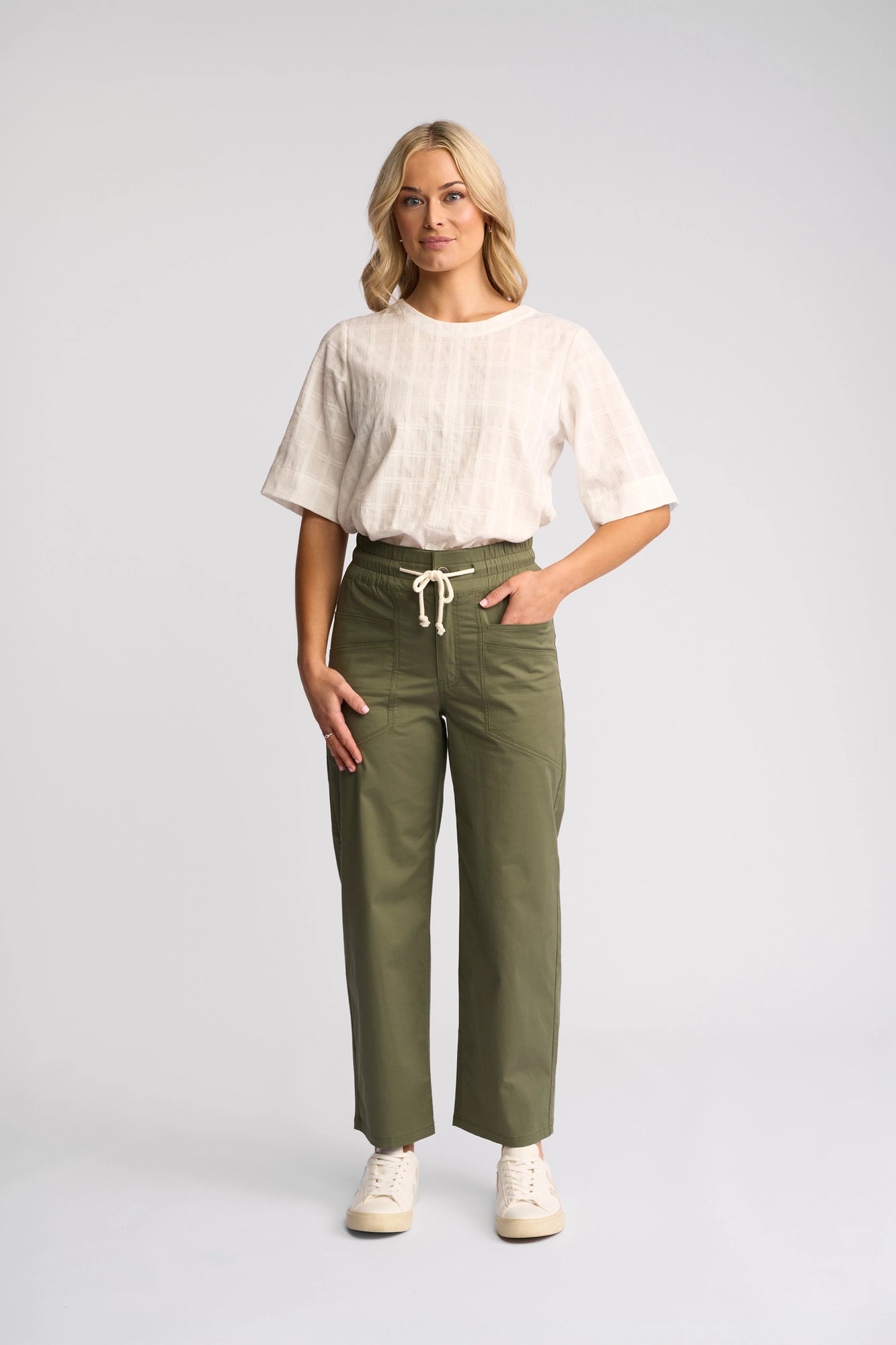 A woman stands facing forward, wearing a white short-sleeve top and the Vassalli Avon Pant in khaki, crafted from stretch cotton. She pairs the outfit with white sneakers. Her wavy, shoulder-length blonde hair frames her face as she maintains a neutral expression against a plain light gray background.