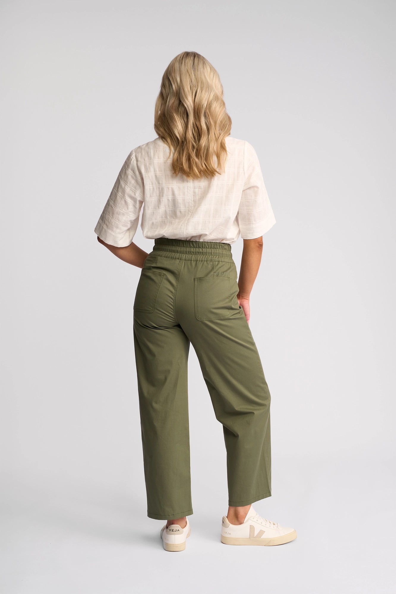 A person with wavy blonde hair stands facing away, wearing a white short-sleeve top, Vassalli Avon Pant - Khaki, and white sneakers against a plain light gray background.