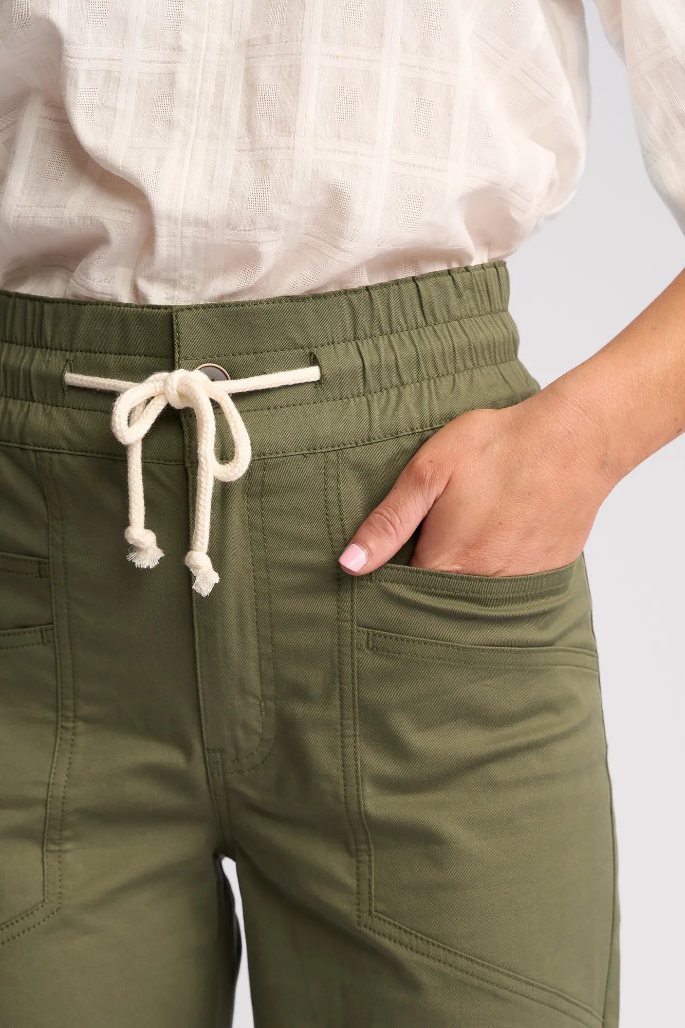 A person wearing Vassalli Avon Pant in khaki, crafted from comfortable stretch cotton with front pockets and a white drawstring, paired with a white textured blouse. One hand is casually placed in the pocket.