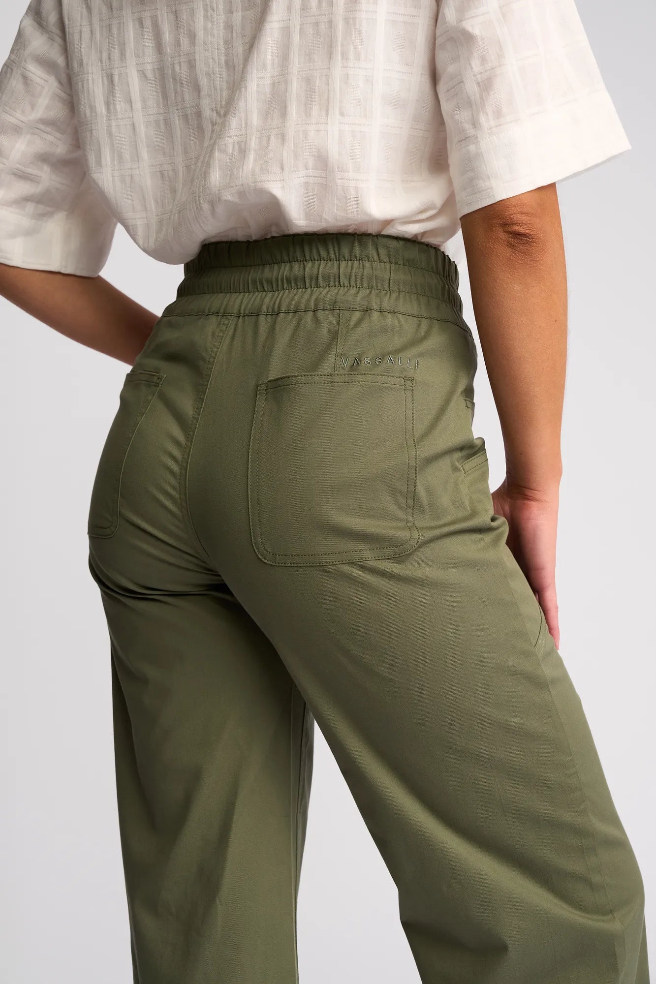 A person wearing an off-white textured top and khaki Vassalli Avon Pant stands with their back to the camera, highlighting the back pocket and elastic waistband detail of the stretch cotton pants.
