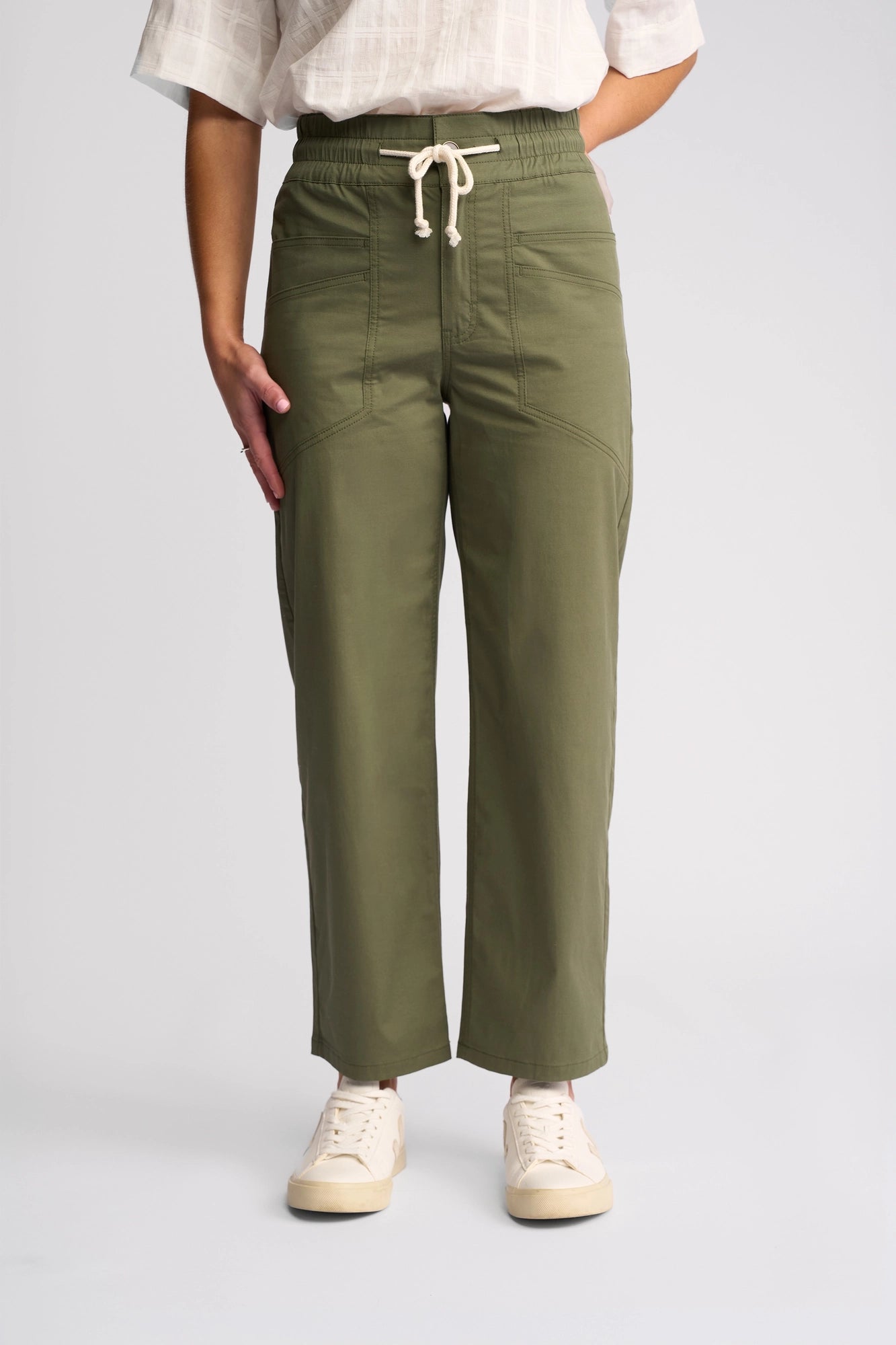 A person is shown from the waist down, wearing the Vassalli Avon Pant in khaki—straight-leg pants made from comfortable stretch cotton with a drawstring waist—paired with a white short-sleeve top and white sneakers, standing against a plain light background.