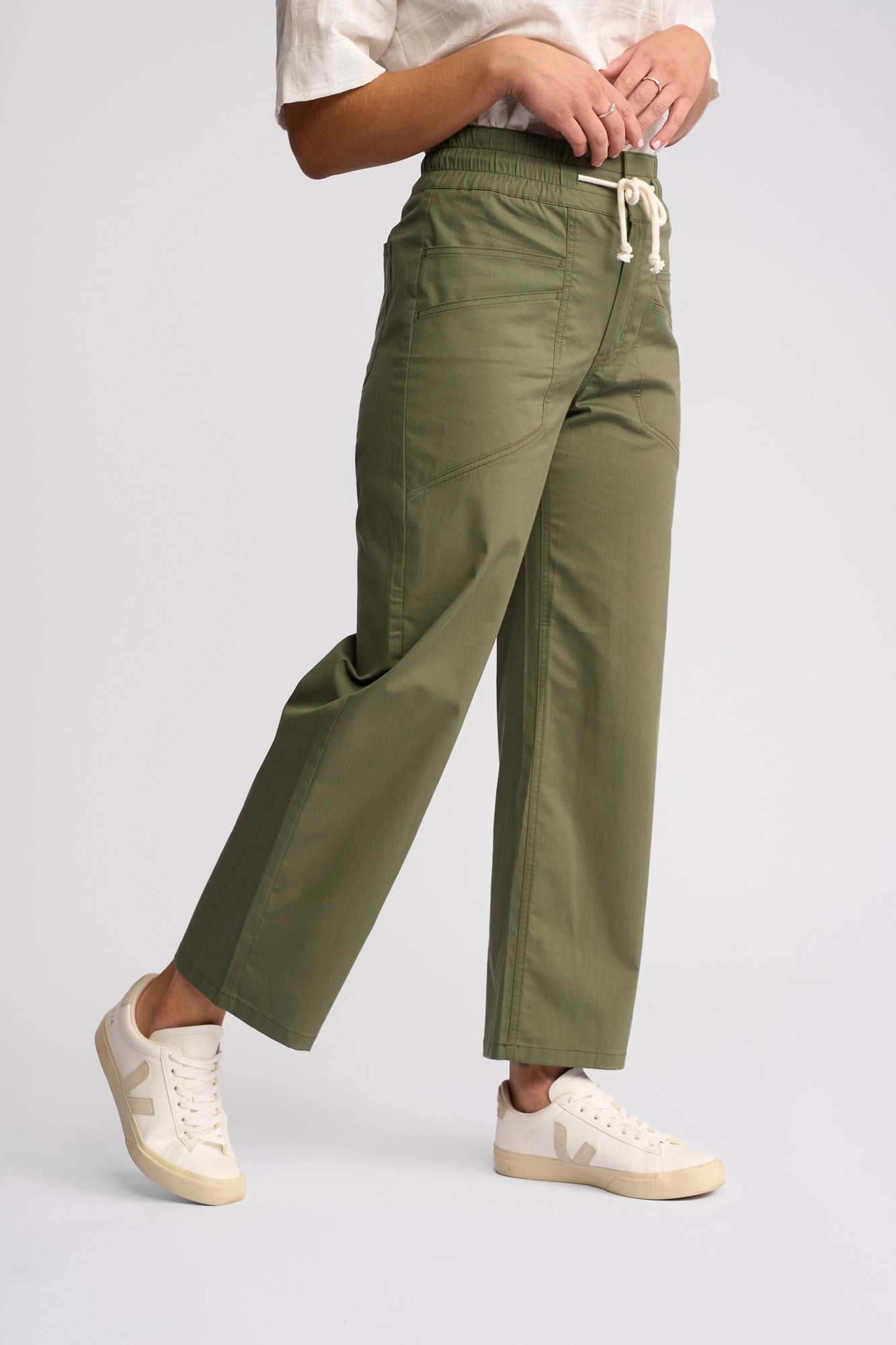 A person is shown from the waist down, highlighting the comfort and casual style of the Vassalli Avon Pant in khaki—a wide-leg silhouette crafted from stretch cotton with a drawstring waist. The look is completed with white sneakers and a white shirt, set against a plain light background.