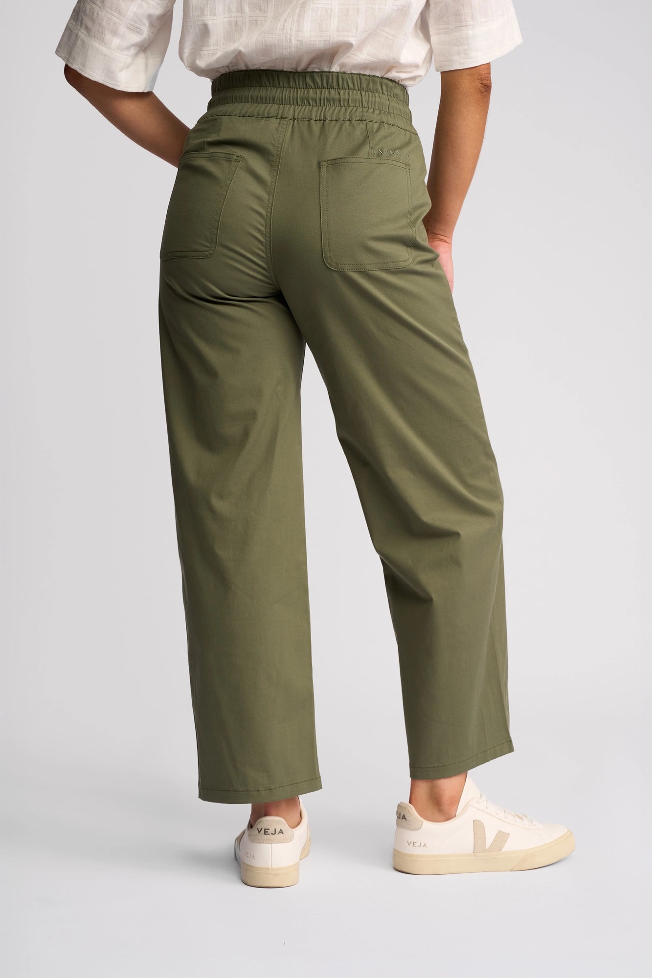 A person stands facing away, wearing the Vassalli Avon Pant in Khaki, paired with a white top and off-white Veja sneakers. The background is plain light gray.