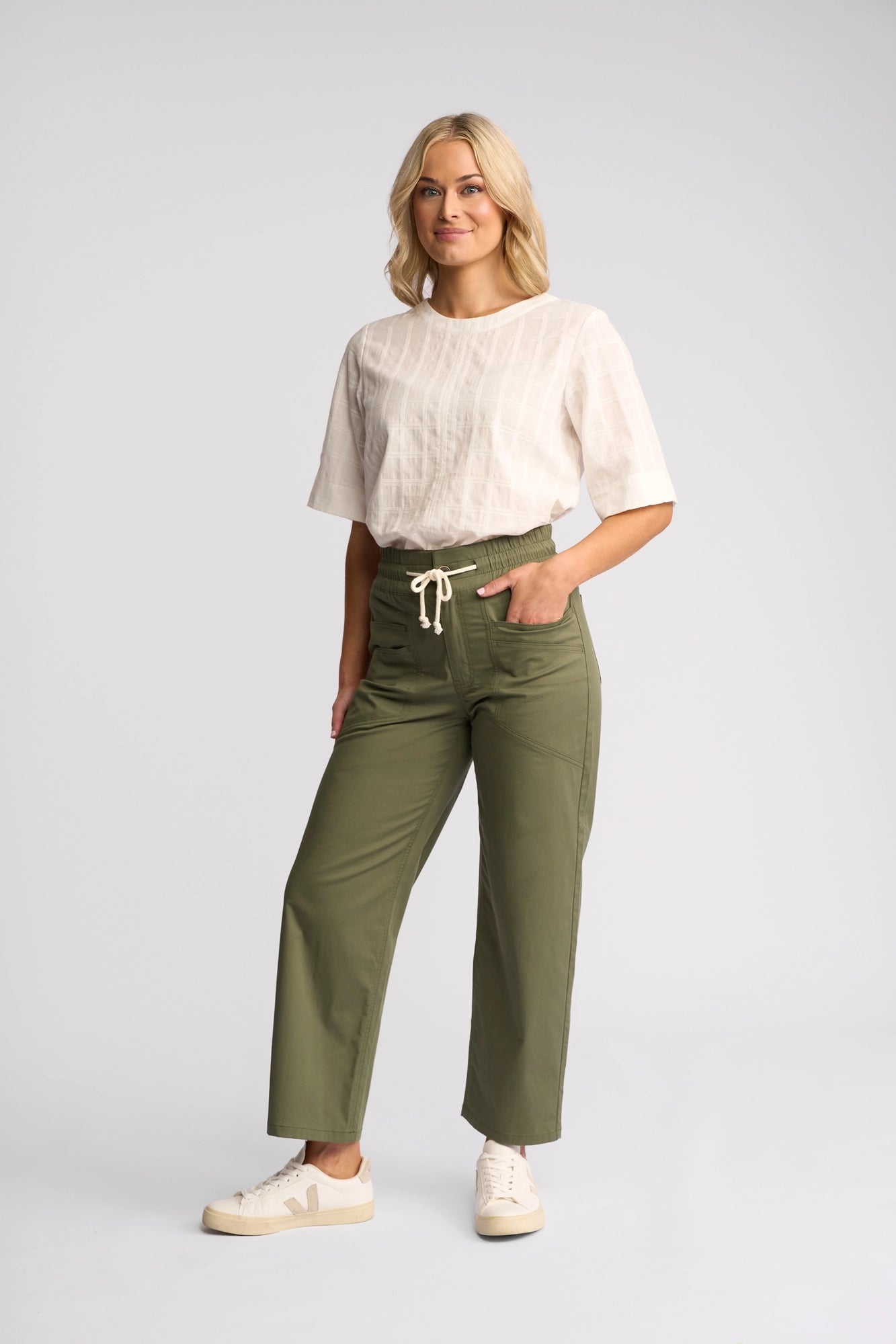 A person stands against a plain background, wearing a white short-sleeve top, Vassalli Avon Pant in khaki crafted from soft stretch cotton, and white sneakers.