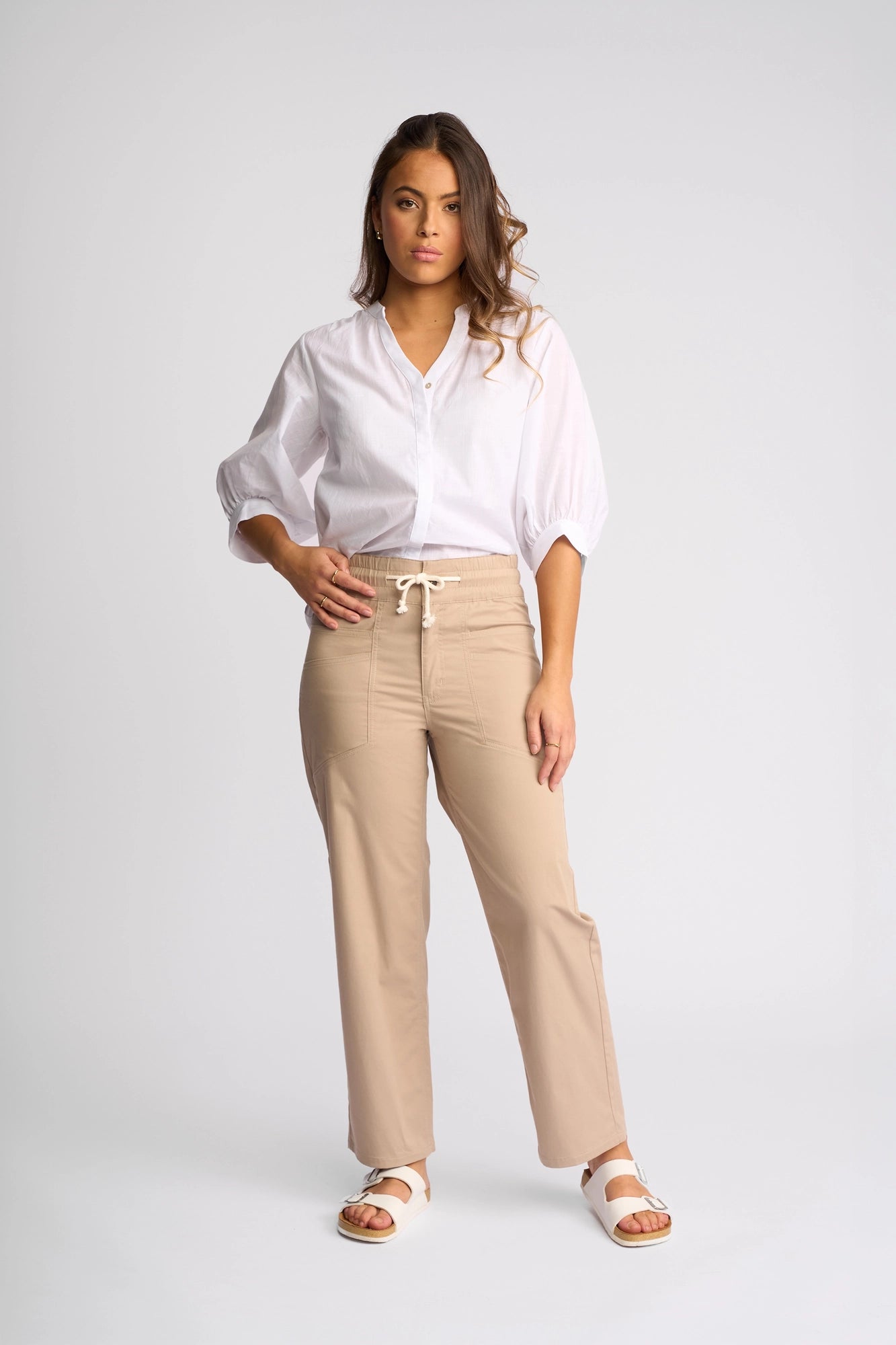 A woman stands facing the camera wearing a white blouse, Vassalli Avon Pant in sand—a beige straight-leg ankle-grazer crafted from stretch cotton—and white sandals against a plain light background, showcasing understated utility-inspired styling.