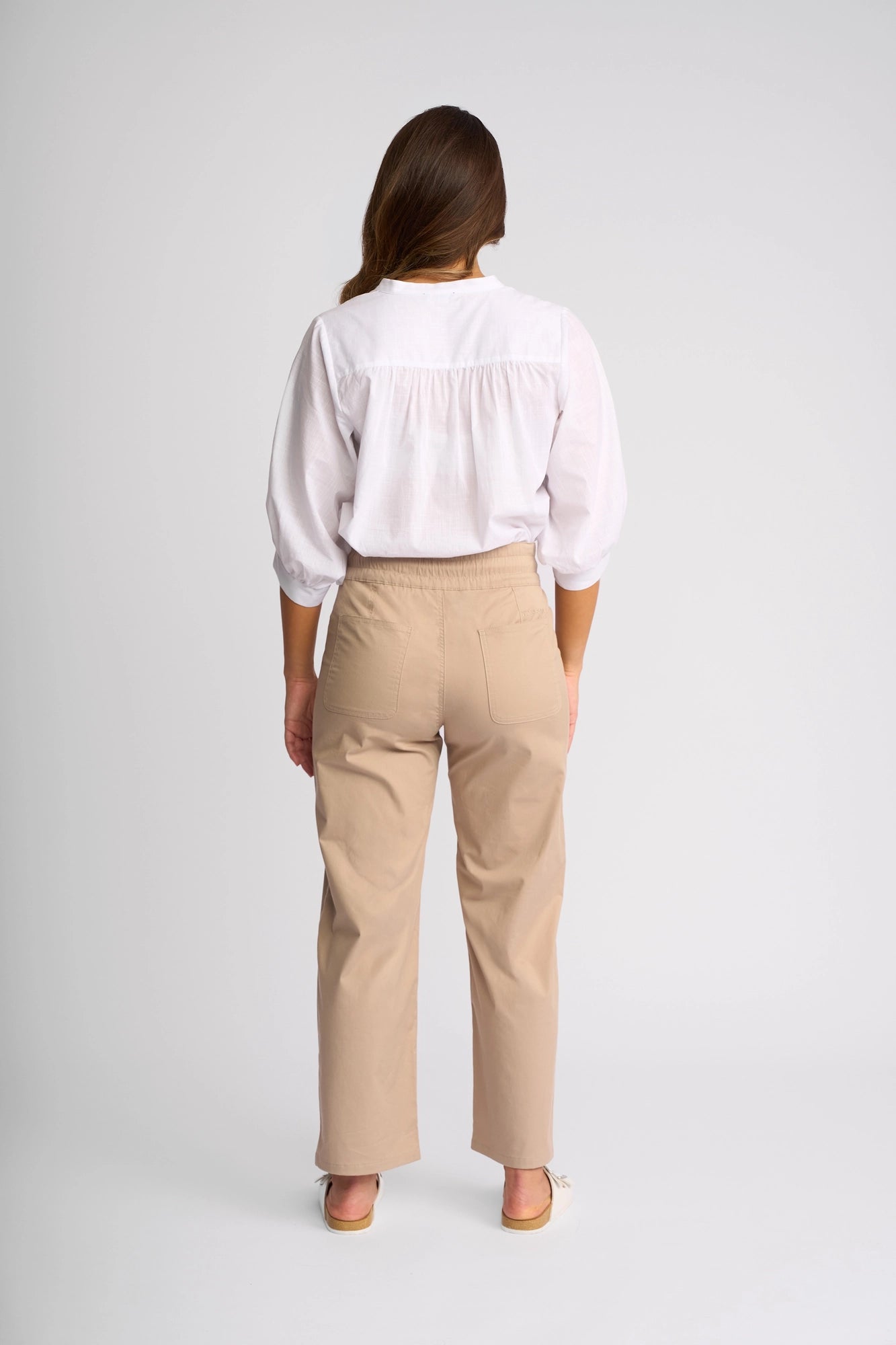 A woman stands facing away, wearing a white blouse with three-quarter sleeves and the Vassalli Avon Pant in Sand—high-waisted, straight-leg ankle-grazer pants crafted from stretch cotton. She is in a plain, well-lit studio setting with a neutral background.